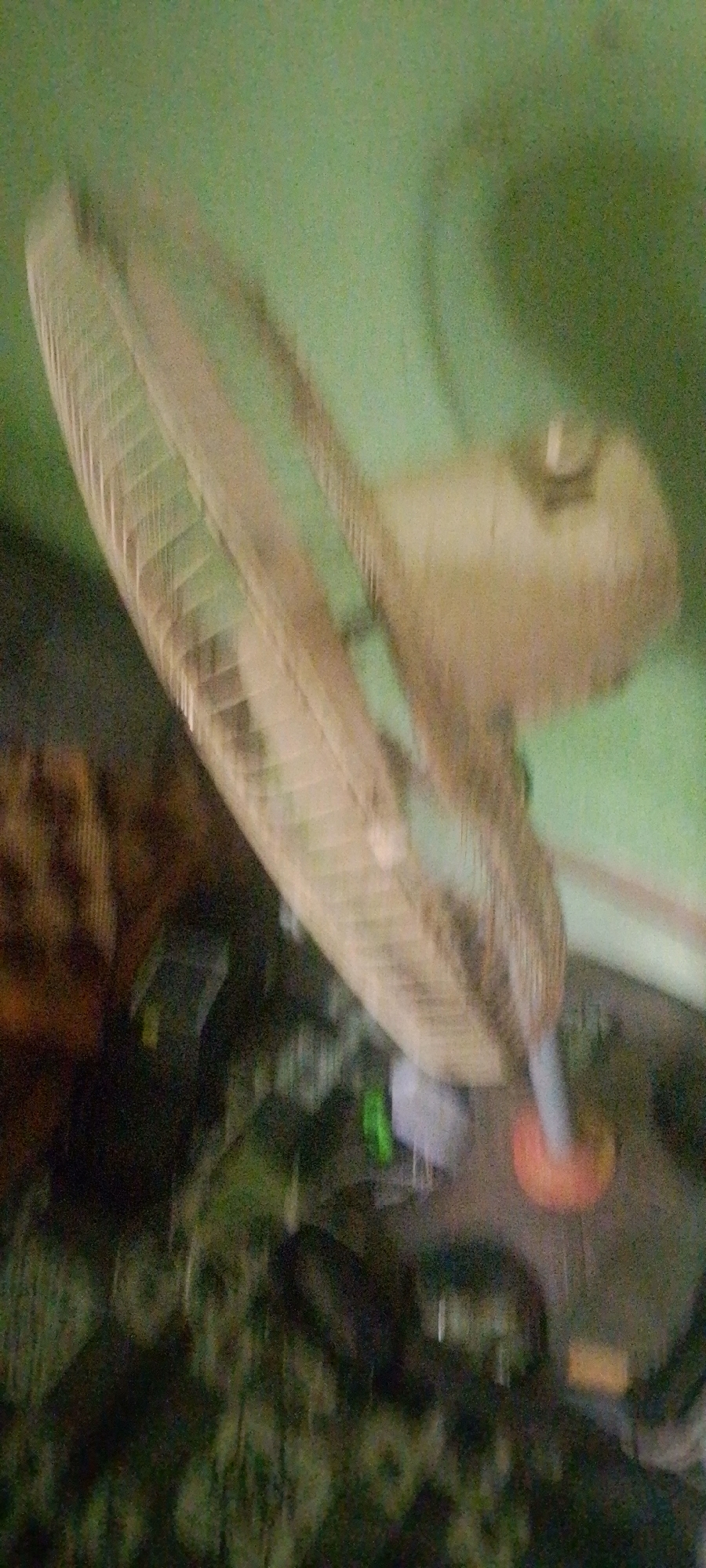 The image shows a close-up of a fan, possibly in an indoor setting with a green wall in the background. The fan appears to be old or dusty, with visible blades and a cage. The image is blurred, suggesting movement or low light conditions. The lower part of the image shows a dark, indistinct background, possibly furniture or other objects. There is a bright orange object visible near the base of the fan, which could be a part of its stand or an unrelated item. The specific location in Jalingo, Nigeria, cannot be determined from the image itself.