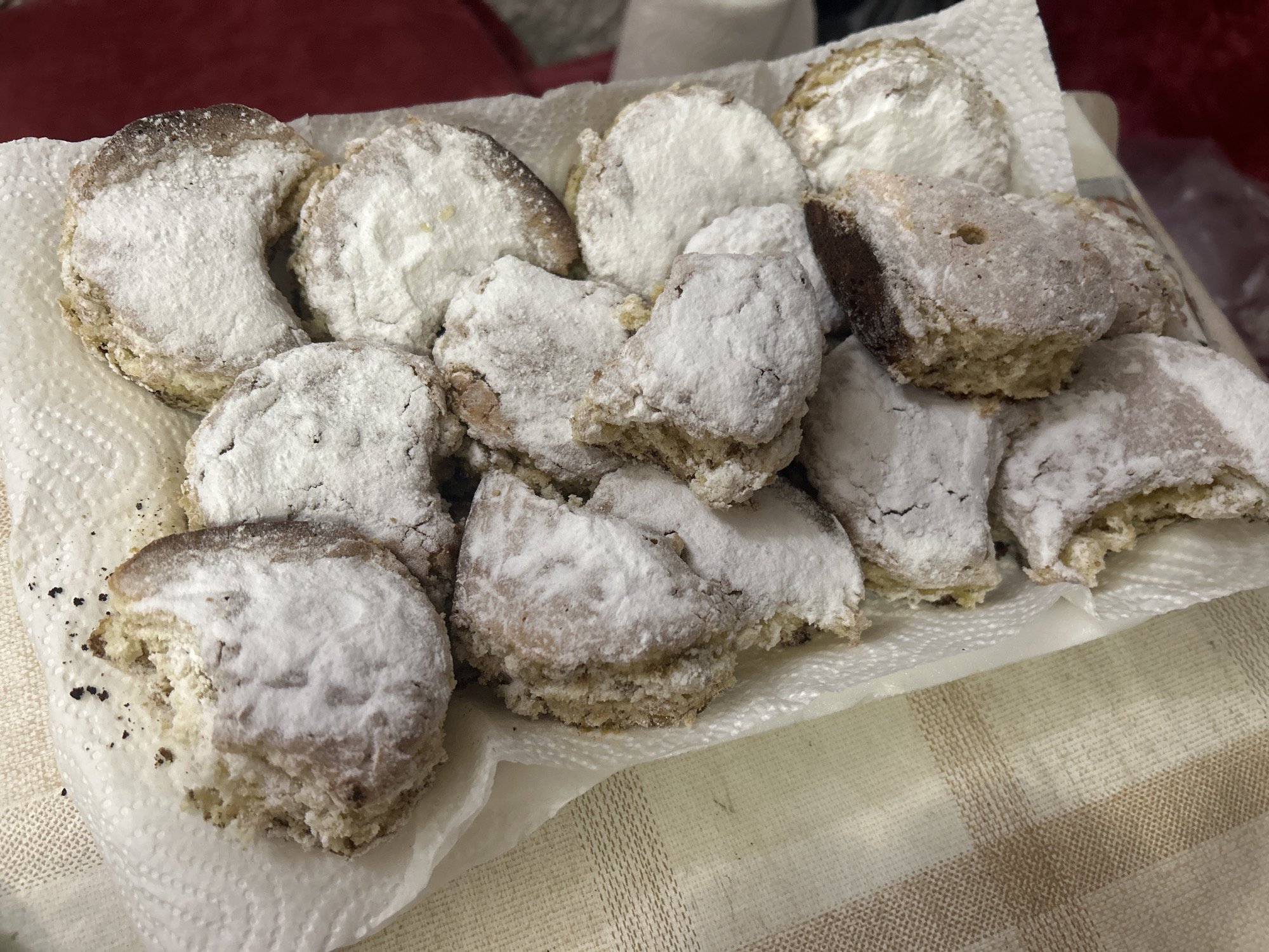 A still life presents a collection of approximately 14 powder-sugared cookies arranged on a white paper towel. The paper towel rests on a tan and white striped tablecloth.  A red cushion is partially visible in the upper left background. The objects are indoors. There are no people visible. The cookies are heavily coated with powdered sugar. The location in Cherven, Bulgaria cannot be inferred from the image.