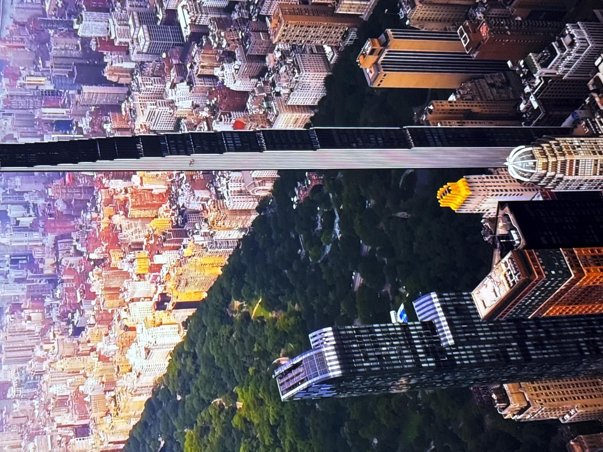 This is an aerial, high-angle, eye-level shot of a dense urban cityscape dominated by a large park and numerous skyscrapers. The park, likely Central Park given the context of similar imagery, occupies the left side of the frame, a vast expanse of dark green trees. Adjacent to the park, to its right, are rows of buildings. The closest building to the foreground on the left is a dark, modern skyscraper with a distinctive stepped crown. Further to the right, a very tall, slender skyscraper with a black and white facade stands prominently in the center of the frame. Surrounding these central structures are numerous other buildings of varying heights and architectural styles, some appearing older and ornate, others more contemporary and boxy. The buildings are a mix of colors, including beige, orange, pink, and white, with many windows reflecting the light. The skyline is immense and stretches into the distance, with the furthest buildings appearing in shades of purple and pink, suggesting either distance or atmospheric conditions. The lighting indicates it may be late afternoon or early evening, with a warm glow illuminating some of the buildings, casting long shadows. There are no discernible people or specific actions captured in the image. The location is not Burgas, Bulgaria, but rather a major metropolitan city, most likely New York City given the presence of Central Park and the characteristic architecture.