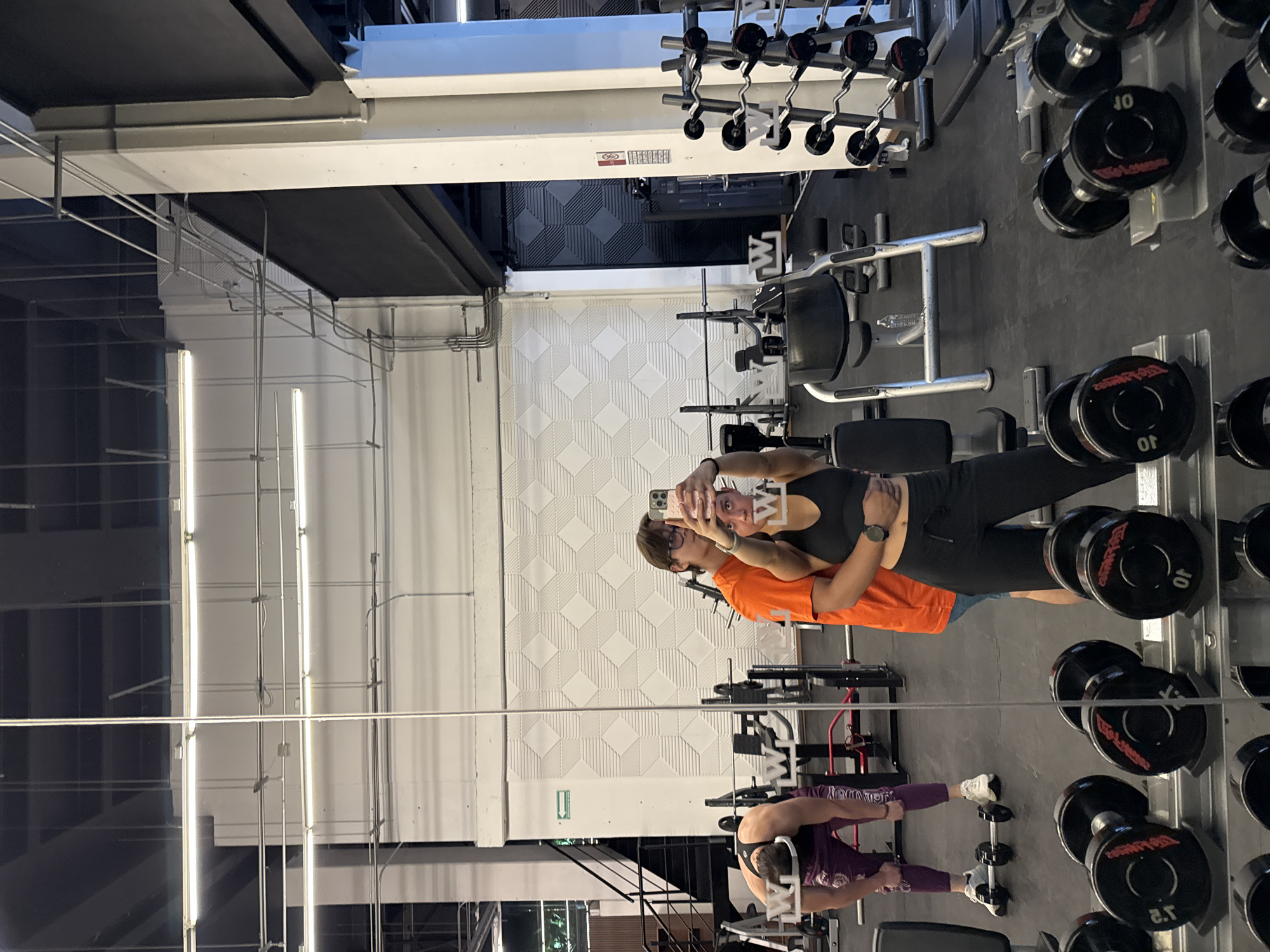 The photograph captures a mirrored scene inside a gym, presumably in Puebla, Mexico given the presence of the gym's logo. Two individuals, one wearing an orange shirt and the other a black sports bra and leggings, are taking a selfie in front of the mirror. Their faces are obscured by the phone they are using for the picture. In the background, another person is visible, wearing purple athletic wear. The gym is equipped with dumbbells of various weights arranged on racks, and weightlifting machines. The walls are white with a geometric pattern. The lighting is provided by fluorescent tubes mounted on the ceiling.