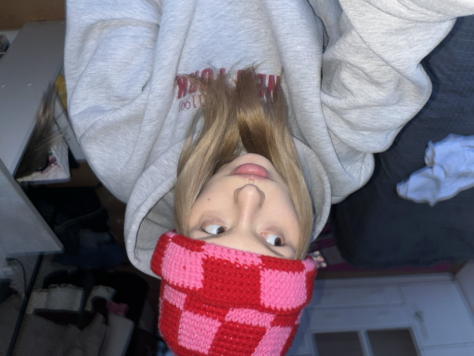 The image captures a close-up, upside-down perspective of a person, likely in an indoor setting. The individual has long, straight blonde hair that falls around their face. They are wearing a light grey hooded sweatshirt, and visible on the front of the sweatshirt, in red lettering, are the words "NEW YORK" and the numbers "1990", though they appear inverted due to the image's orientation.
The person is also wearing a prominent red and pink checkered knit beanie or hat, which covers their forehead. Their face is positioned at the bottom of the frame, with their head tilted back, looking upwards towards the camera. Their eyes are slightly rolled to the side, giving them a playful or quizzical expression. Their mouth is slightly open, and the tip of their tongue is visible.
In the background, to the left of the person (when viewed right-side up), there appears to be a white shelving unit or furniture with various items on it, suggesting a casual, possibly cluttered, personal space. To the right, a dark, solid surface, likely a bed with dark bedding, is visible, and a crumpled white item of clothing rests on it. The overall impression is a casual, relaxed moment captured from an unconventional angle.