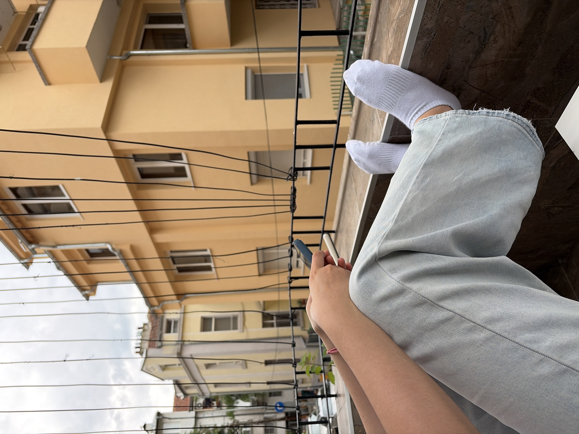 A low-angle, medium shot depicts a person sitting, wearing white socks and light-wash jeans. The person is holding a blue smartphone and appears to be interacting with it. In the background, there is an exterior view of a building with light yellow facade and visible windows. Thin wires run across the field of view, partially obscuring the building. The photograph appears to be taken from a balcony, and a fence is visible. Based on visual similarities to architecture in Burgas, Bulgaria, the location could be Burgas, Bulgaria.