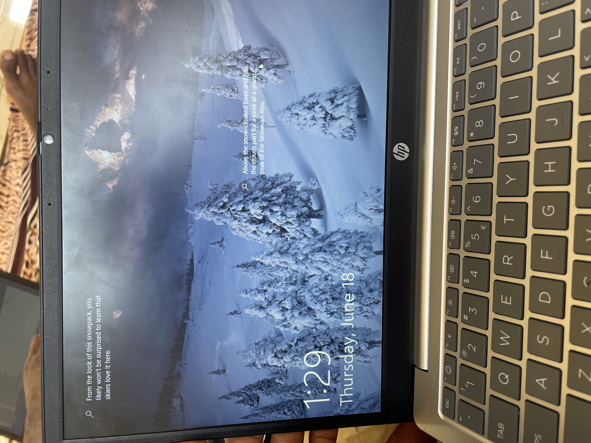 The image displays the lock screen of an HP laptop. The background is a serene winter landscape with snow-covered pine trees and mountains under a cloudy sky. The time "1:29" is prominently displayed in the bottom left corner, followed by "Thursday, June 18". To the left of the time, a search icon precedes the text "From the look of this snowpack, you likely won't be surprised to learn that skiers love it here." On the right side of the screen, another search icon is present, followed by the text "Above the snow-cloaked trees and hills, the clouds part for a peek at a grand peak of the Slovenian Alps." The HP logo is visible on the laptop's bezel below the screen. The keyboard of the laptop is also visible in the foreground. The image is a close-up of the laptop screen and keyboard, taken at an angle. There is no information in the image that suggests the location is Wai A, Nigeria.