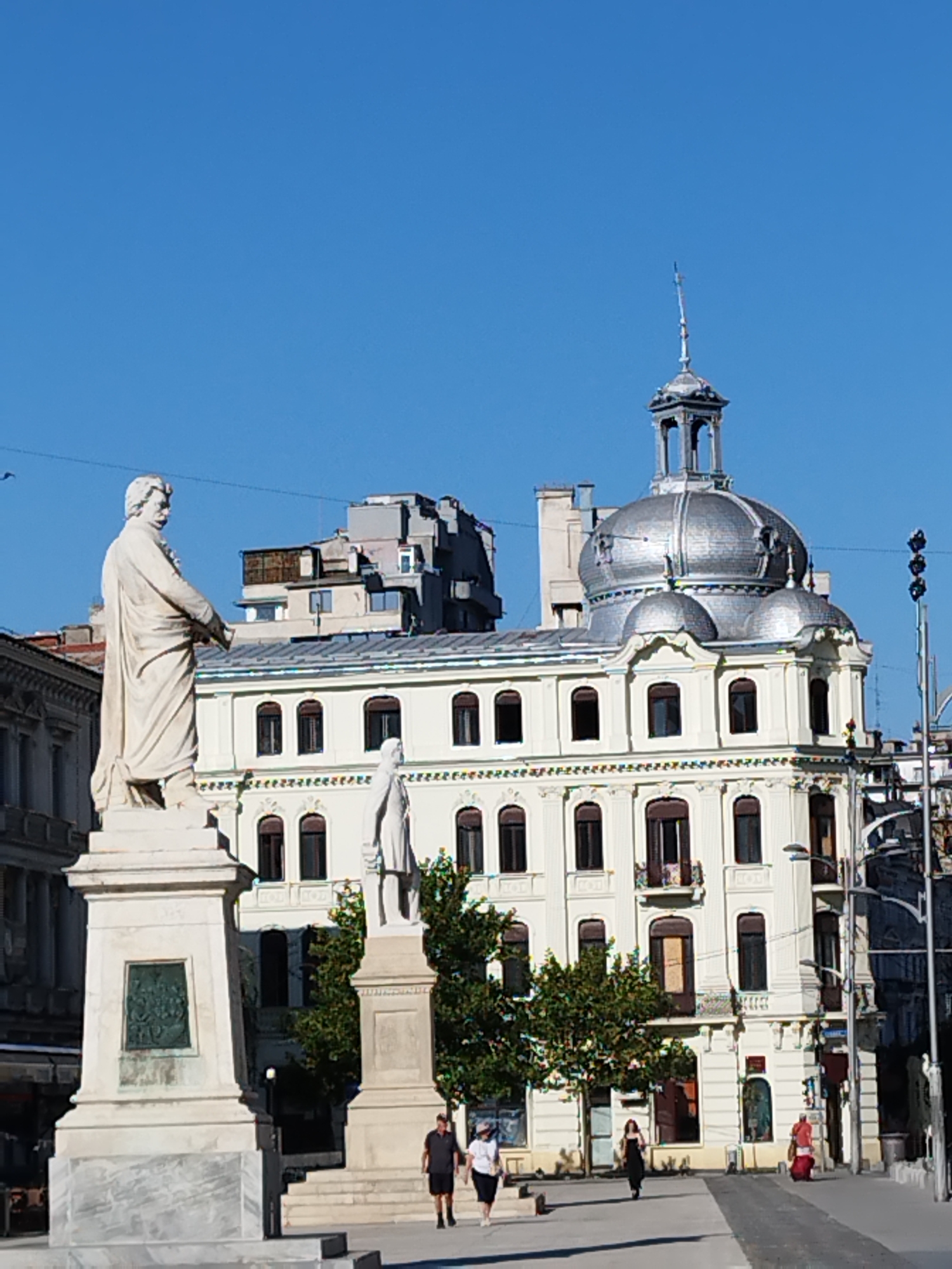 The image depicts a public square in Bucharest, Romania, dominated by a large white stone statue of a man in historical attire on the left. The statue stands on a tiered pedestal, which features a bronze plaque with relief imagery on its front. To the right of this statue, a smaller white stone statue of a robed figure is visible.
Behind the statues, a grand, pale yellow building with ornate white trim and dark-capped windows is prominent. The building features a distinctive silver dome with a small cupola topped by a spire. Other architectural elements include arched windows, balconies, and decorative moldings. Several trees with lush green foliage are situated in front of the building.
The sky is a clear, bright blue. In the foreground, a paved walkway is visible, with several people walking. On the left, two individuals, a man in a dark shirt and shorts and a woman in a white shirt and dark pants, are walking away from the statues. Further to the right, a woman in a black dress is walking towards the viewer, and another person in a red garment is seen walking away on the right side of the frame. The ground is a mix of pavement and what appears to be a grey stone surface. The scene suggests a sunny day in a city center, possibly a historical or cultural district.