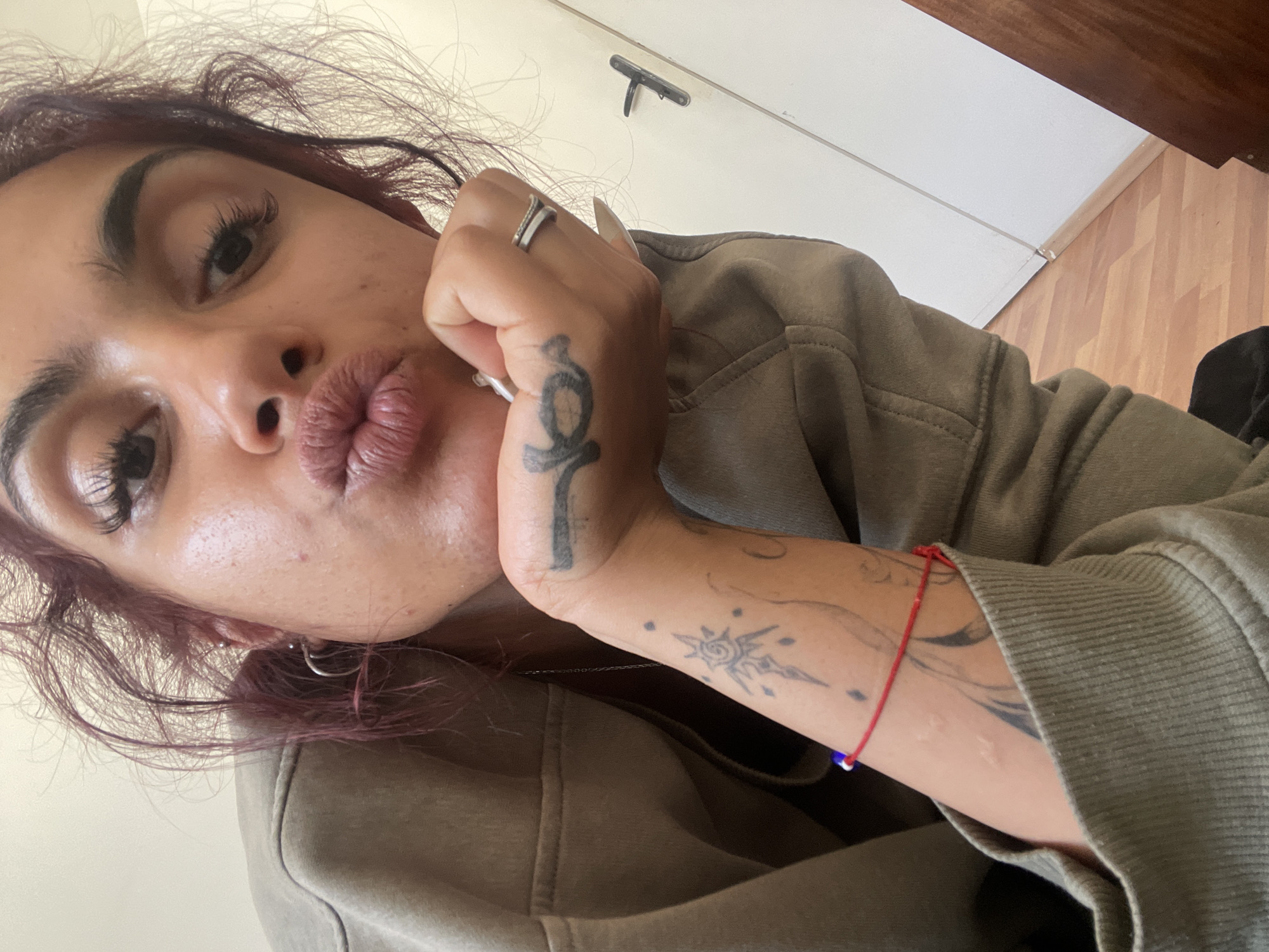 The image presents a low-angle, close-up self-portrait of an adult individual located in Unknown, Unknown. The subject has medium-dark skin, dark reddish-brown curly hair, and is looking directly at the camera with lips pursed in a "kissing" expression. Long, dark eyelashes and thick eyebrows are prominent. The subject's chin rests on their right hand, which has long, light-colored fingernails and a two-band silver-colored ring on the middle finger. A small dark tattoo, resembling a stylized sword or dagger, is present on the back of the right hand. The individual wears a light olive-green hooded sweatshirt. The visible right ear has at least two piercings: a silver hoop and a stud. On the inner left forearm, a tattoo of a stylized sun or starburst with radiating lines and dots is visible, along with scattered dots and a portion of a larger, abstract dark tattoo. A thin red string bracelet with a small blue bead is worn on the left wrist. The background features light brown wooden floorboards and a plain white wall. A white door with a dark, flat-handled lever is partially visible on the right side of the frame.