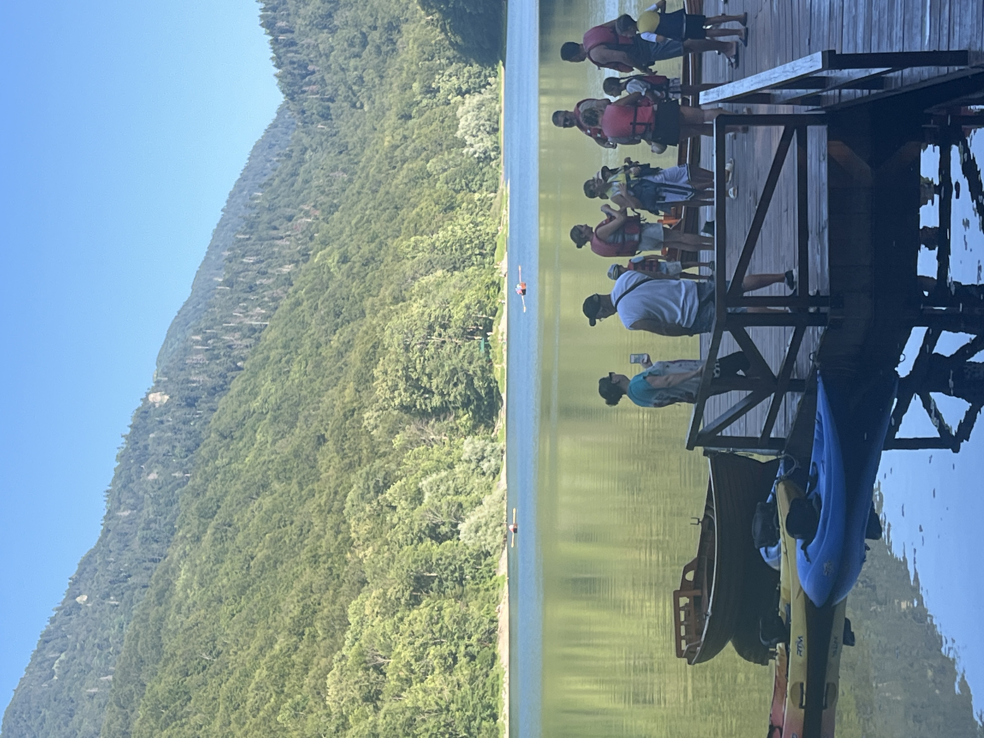 This image depicts a serene lake scene in Sjerogošte, Montenegro. A group of people, including adults and children, are gathered on a wooden pier. Many are wearing life jackets, suggesting they are preparing for or have just finished water activities. Several kayaks and a rowboat are docked at the pier, with reflections visible in the calm water. In the background, a vast, tree-covered mountain range rises under a clear blue sky. The lake's surface is smooth, mirroring the surrounding lush greenery and the sky. The overall atmosphere is peaceful and suggests a recreational setting.