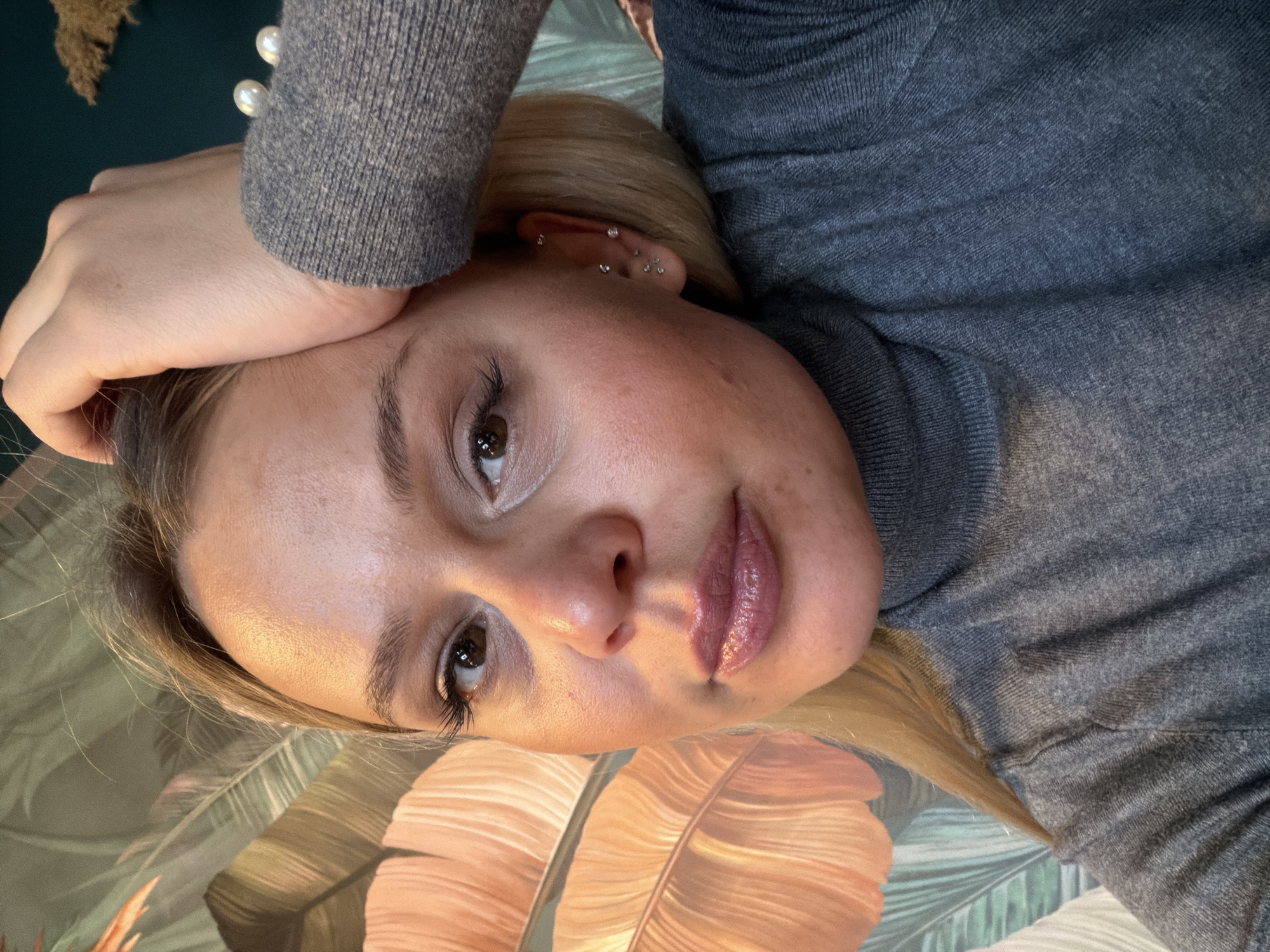 A woman with blonde hair is lying on a surface with a leaf pattern. She is wearing a grey turtleneck sweater with two pearl buttons on the sleeve. Her left hand is resting on her forehead. She has multiple piercings on her left ear. There is no information to ascertain a location in the city of Sarapul, Russia.