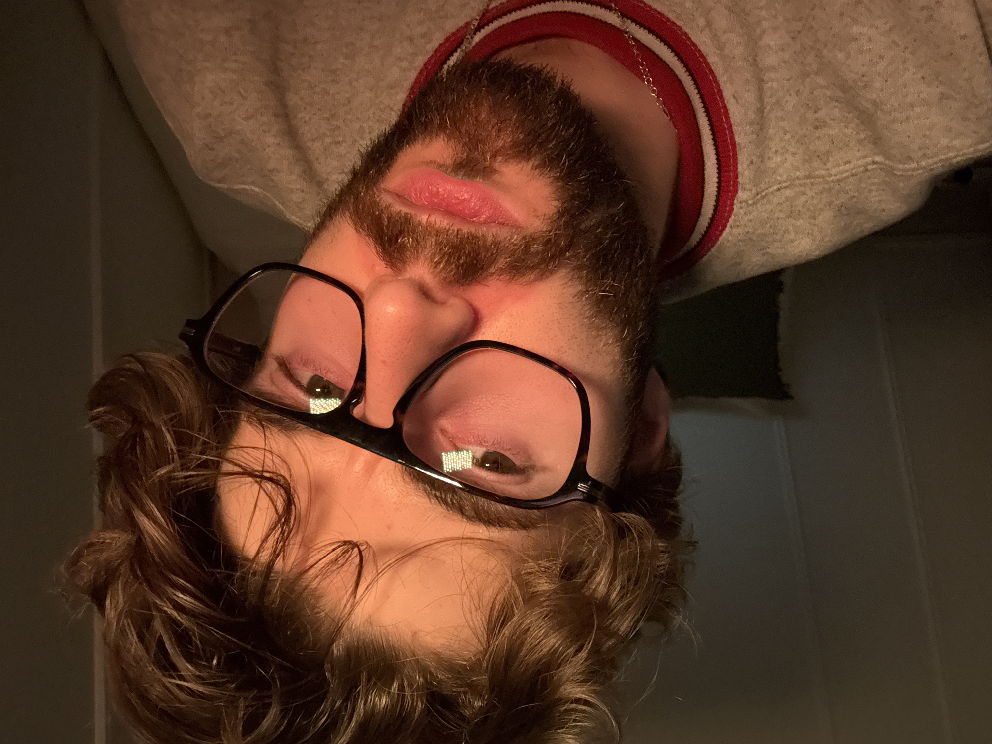 A selfie taken by a bearded person wearing glasses and a grey sweater with a red and white collar. The person is upside down in the frame. The background appears to be an interior space with a dark wall and a glimpse of what might be a doorway. The location in the city of Montreal, Canada is unknown.