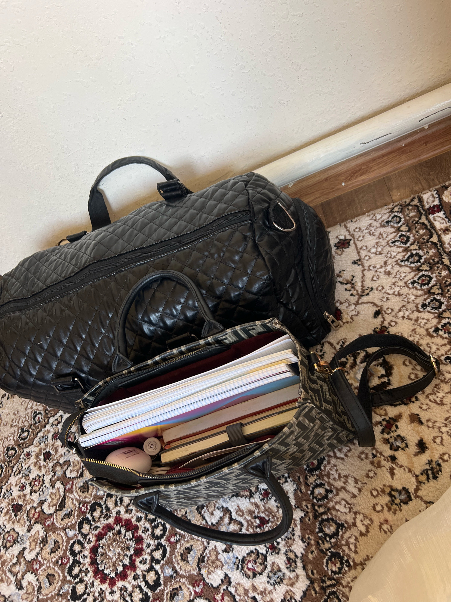 The image shows two black duffel bags and a smaller patterned handbag on a patterned rug. One duffel bag is quilted and larger than the other. The smaller handbag is open, revealing several notebooks, a cylindrical object with a pink cap, and a round, pale object. The bags and handbag are situated on a rug with a red and beige floral pattern. A white wall and a wooden floor with a baseboard are visible in the background. The scene appears to be indoors, possibly in a residential setting. There are no people or animals visible in the image. The lighting suggests it is daytime. There is no indication of the location in Vinogradnoye, Kyrgyzstan.
