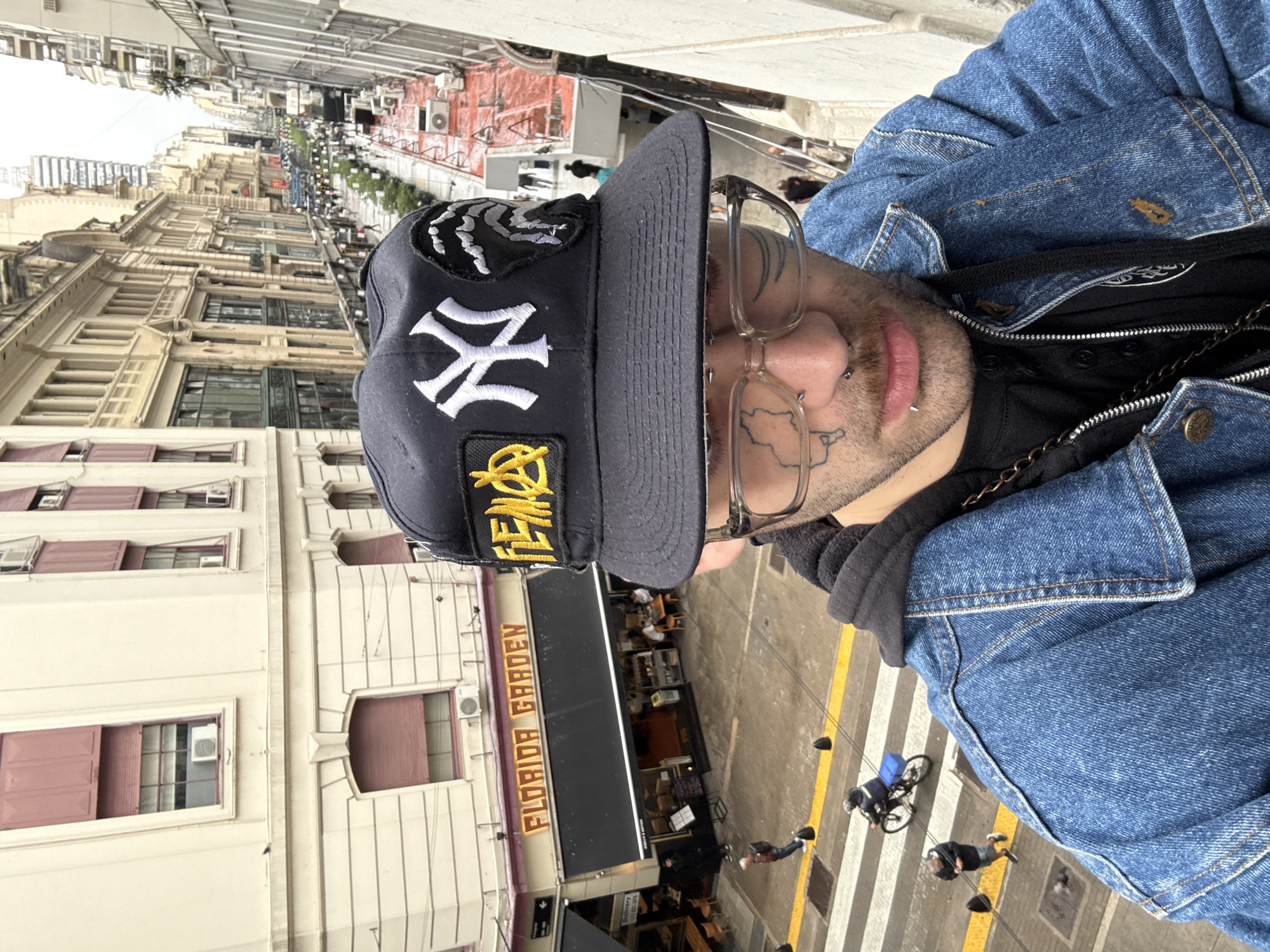 The image, taken from an elevated viewpoint, features a person looking downwards into a bustling city street. The individual, seen from the chest up, wears a blue denim jacket over a black hooded top. They have clear-framed glasses, facial piercings (including septum and philtrum), and visible tattoos on their cheek and temple. A black baseball cap adorns their head, featuring a prominent white "NY" logo (New York Yankees), a yellow patch with a symbol and the word "TEMPO," and a textured black-and-white patch on the side.
Below the person, an urban landscape unfolds, characteristic of a major city like Buenos Aires, Argentina. Multi-story buildings with classical architectural details and numerous windows line the street. Many windows have closed reddish-brown shutters, and air conditioning units are visible on building exteriors. Directly beneath the viewpoint, a business with a dark awning displays "FLORIDA GARDEN" in gold lettering. The street itself shows pedestrians walking and a person on a bicycle with a blue delivery box, traversing a clearly marked crosswalk. The scene is illuminated by daylight, suggesting an overcast sky due to the even, diffused lighting.