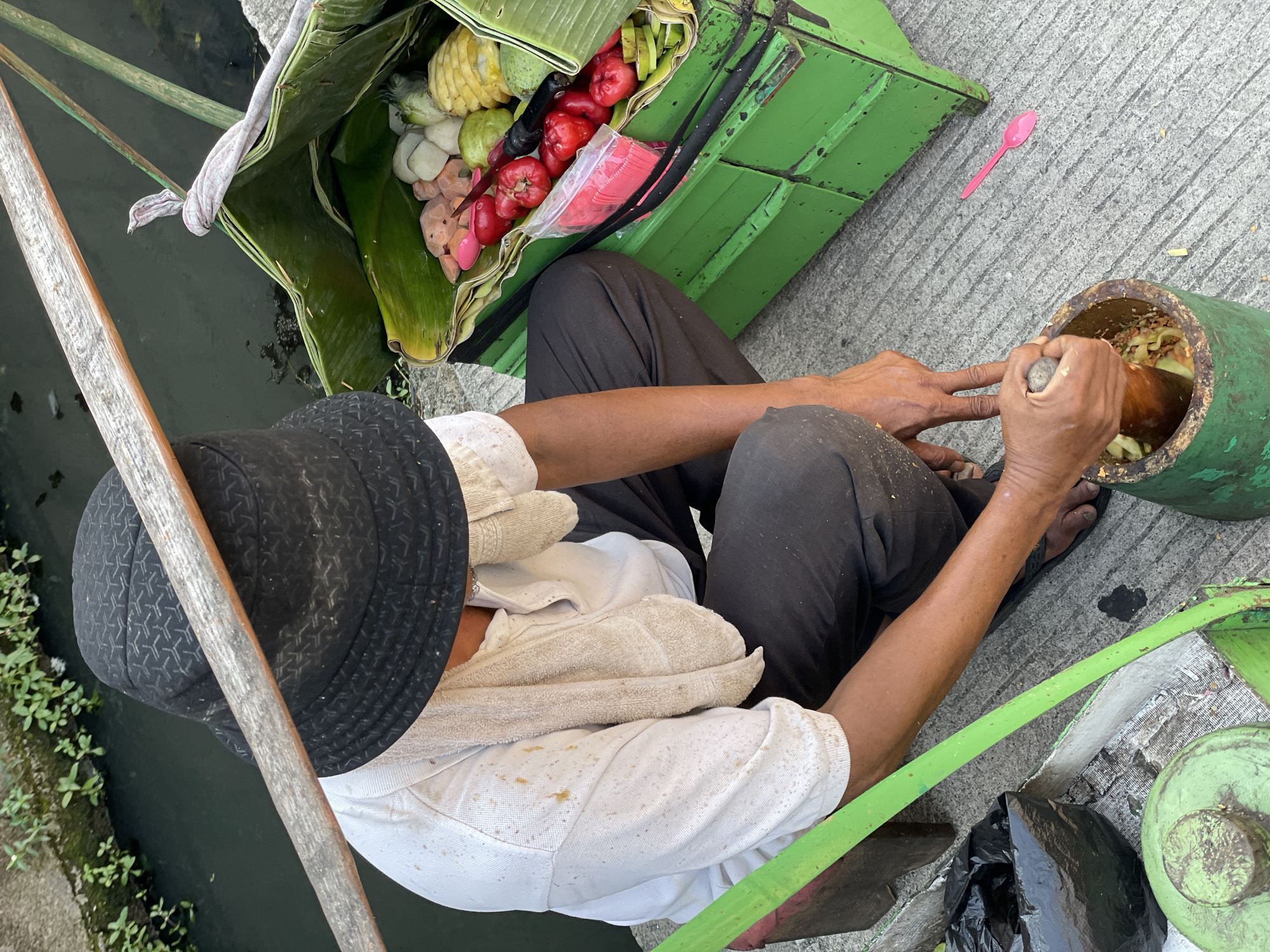 A street vendor is shown from a high-angle perspective in North Jakarta, Indonesia. He is crouched beside a narrow canal, preparing fruit salad. The vendor wears a black hat, a light-colored, stained shirt, and dark pants. He sits beside his green vendor cart, which holds a variety of fruits arranged in banana leaves, including sliced pineapple, guava, rose apples, and other unidentified fruits. He is using a wooden pestle to mix ingredients in a green mortar. A pink plastic spoon lies on the concrete beside the cart. The canal is visible to the left, with green plants growing along its edge.