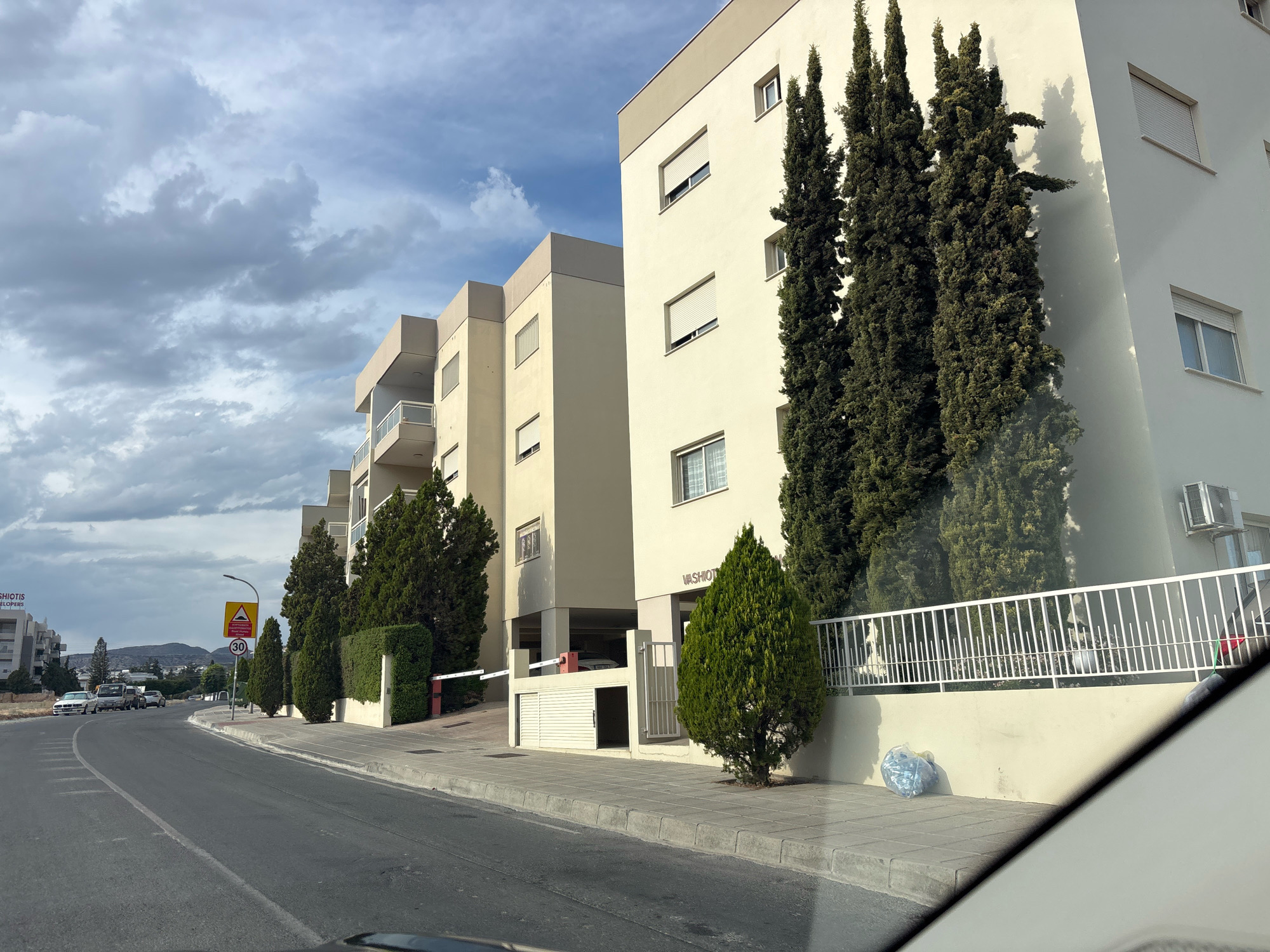 The image depicts a street view in Mesa Geitonia, Cyprus, featuring a row of modern, multi-story apartment buildings. The buildings are light-colored, with prominent balconies and numerous windows, some equipped with roll-down shutters. Tall, slender cypress trees are strategically planted along the sidewalk, casting shadows on the building facades. A white metal fence separates the property from the sidewalk.
In the foreground, a paved road curves to the left, with a marked pedestrian sidewalk adjacent to it. Several cars are visible on the road and parked near the buildings. Traffic signs, including a speed limit sign indicating "30", are present along the roadside. One building displays the partial text "VASHIOTE" on its facade. A white boom gate and a small, open garage-like structure with a door are visible in front of one of the buildings.
The sky is partly cloudy, with dramatic grey clouds dominating the upper portion of the frame. The overall impression is a suburban or residential area in Cyprus on an overcast day.