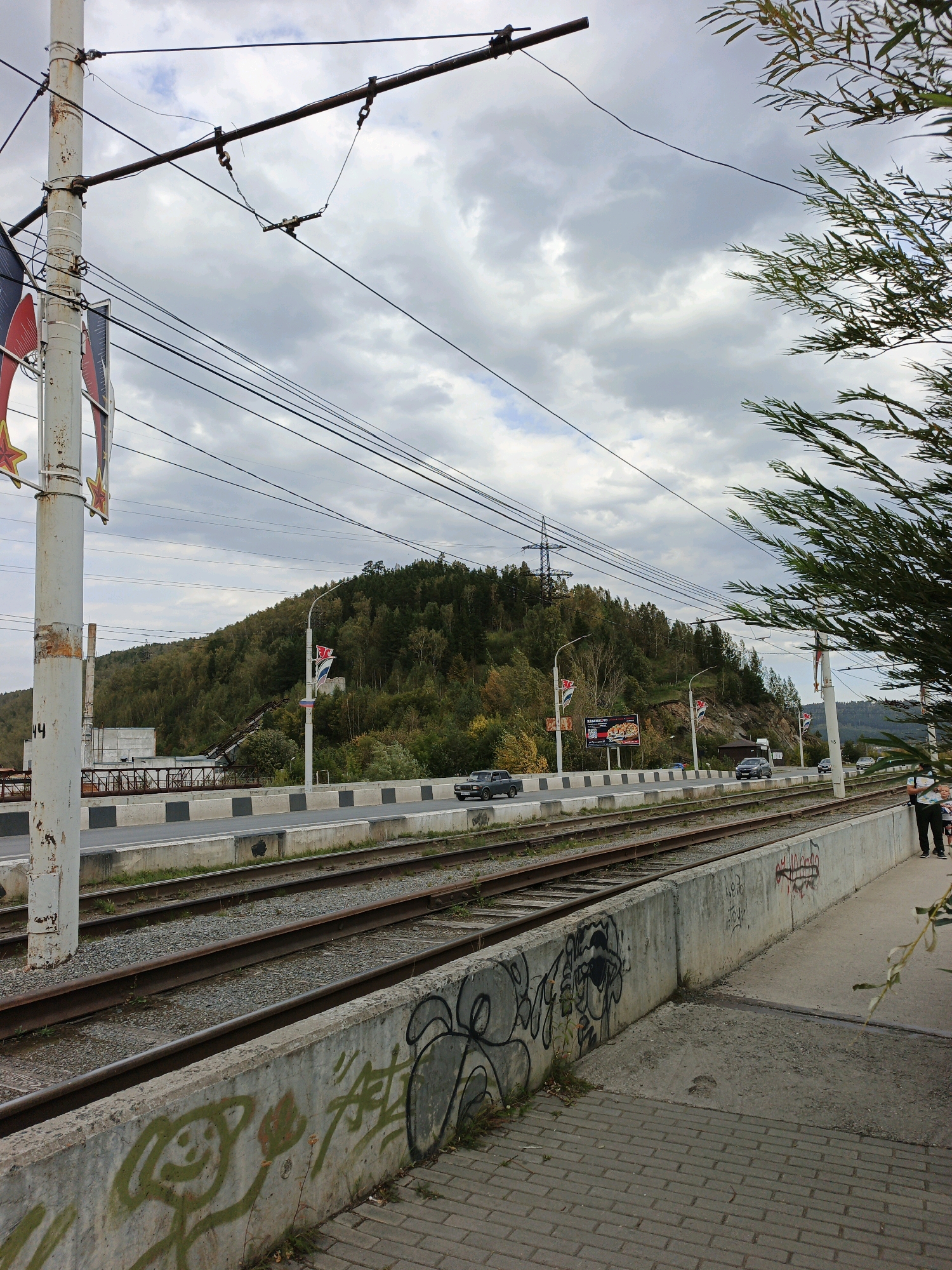 The image shows a cloudy day with a view of a road and train tracks in Zlatoust, Russia. The road is elevated and has several cars driving on it, with a forested hill in the background. The train tracks run parallel to the road, with a concrete barrier between them. The barrier is covered in graffiti. A utility pole with overhead wires is visible on the left side of the image, and a tree with green leaves is on the right. Two people, a man and a child, are standing near the right edge of the image, looking towards the left.
