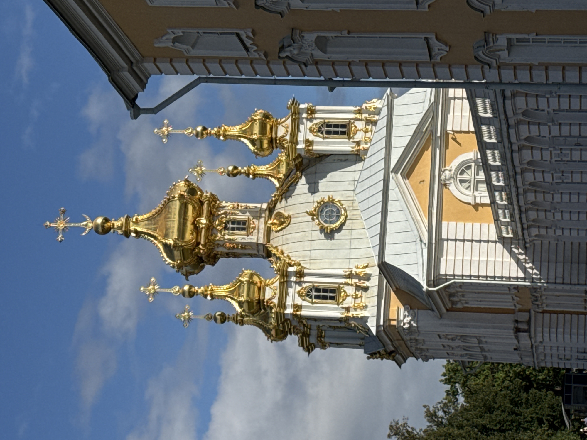 The image shows a detailed view of the Church of the Icon of the Mother of God "The Burning Bush" in Peterhof, Russia. The church features multiple golden domes topped with crosses, set against a backdrop of a partly cloudy blue sky. The building's facade is predominantly white with ornate gold decorations and numerous windows. Adjacent to the church, a yellow building with white architectural details is partially visible. A section of a white balustrade and an arched colonnade is also in view, suggesting a courtyard or promenade area. Green trees are visible on the left side of the image, and the sun casts shadows, indicating it is daytime. No people are clearly discernible in the frame, though faint figures might be present at the very bottom edge. The architecture is characteristic of Baroque style, commonly found in Peterhof's palaces and ensembles.