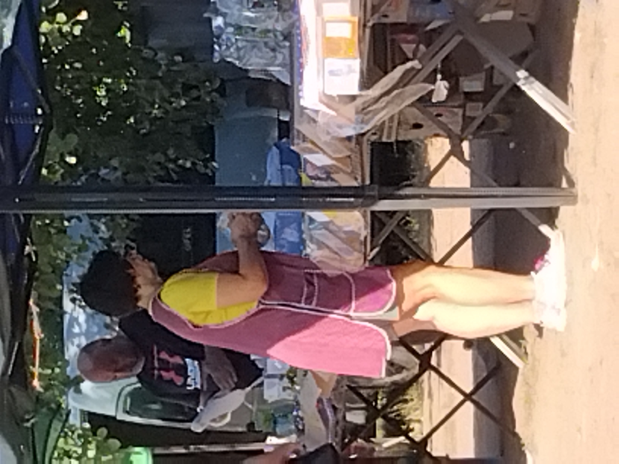 The image captures an outdoor market scene. A woman wearing a pink apron over a yellow shirt and grey shorts stands on the right side, facing left. Her legs and feet are visible, clad in white sneakers with pink accents. She appears to be handling items on a table. To her left, a man in a black t-shirt with a red logo is visible, holding a white paper. Behind them, a white vehicle and foliage are present. The ground is covered in sand or dirt. The market appears to be set up with tables displaying various items, some of which are covered in plastic or bags. The lighting suggests it is daytime. The image does not provide any specific identifiers for the location within Irshansk, Ukraine, beyond it being an outdoor market.