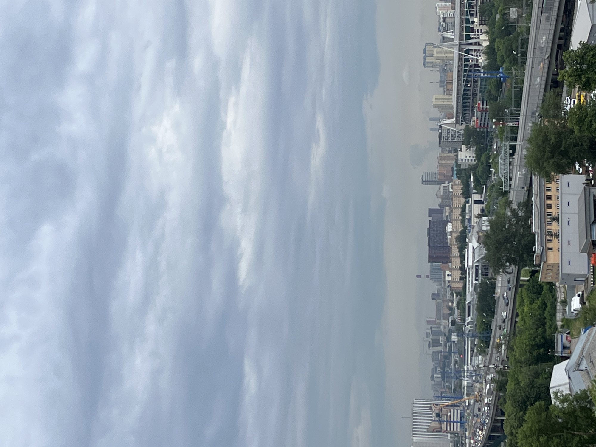 This is a vertical, high-angle, full shot of a city under a cloudy sky. The sky is filled with a thick layer of gray clouds, obscuring the sun and creating a diffused, dim light across the entire scene. In the foreground, the roof of a building with a white and gray facade is visible, with some surrounding greenery and other industrial-looking buildings with white roofs and gray walls.
The midground is dominated by a multi-level highway system with moderate traffic, including cars and trucks. The highway is surrounded by lush green trees. Beyond the highway, a sprawling urban landscape unfolds, featuring a variety of buildings ranging from low-rise structures to tall, modern skyscrapers. A large stadium with white, arching supports is prominently visible in the right midground. Industrial buildings and smokestacks can be seen throughout the city. The overall impression is that of a densely populated, active city.
The image appears to be a cityscape of Tarasovka, Russia, judging by the distinct architecture and the presence of what looks like the Arena CSKA stadium.