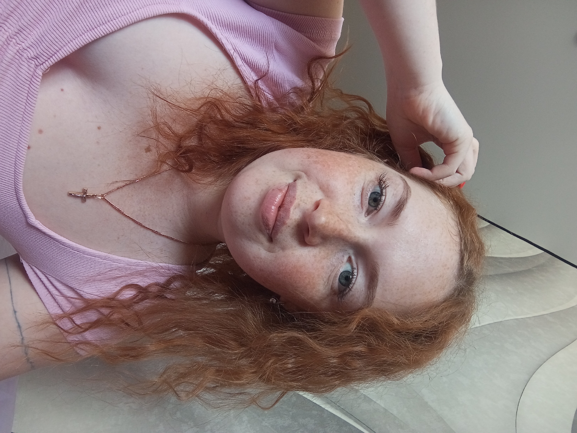 A fair-skinned woman with red, curly hair is positioned at an angle against a light-colored surface with a subtle, curved design. Her face is directed towards the camera, exhibiting freckles and blue eyes. She is wearing a light pink top with a scoop neckline and a delicate gold necklace with a cross pendant. Her right hand is raised near her head. Part of her upper chest and the upper portion of her left arm, which appears to have a tattoo, are visible. The context of the image suggests the scene is indoors. The location is not discernible based on the image alone, beyond that it may originate from Yurgamysh, Russia.