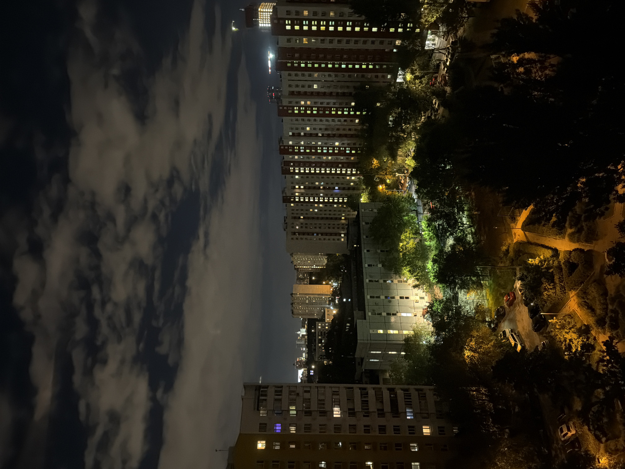 The image presents an elevated view of Khimki, Russia, at night. Tall apartment buildings with illuminated windows dominate the skyline. The left side of the frame shows a cloudy night sky. Below, trees and a street with parked cars are visible, illuminated by streetlights and building lights. The scene suggests an urban residential area at night.