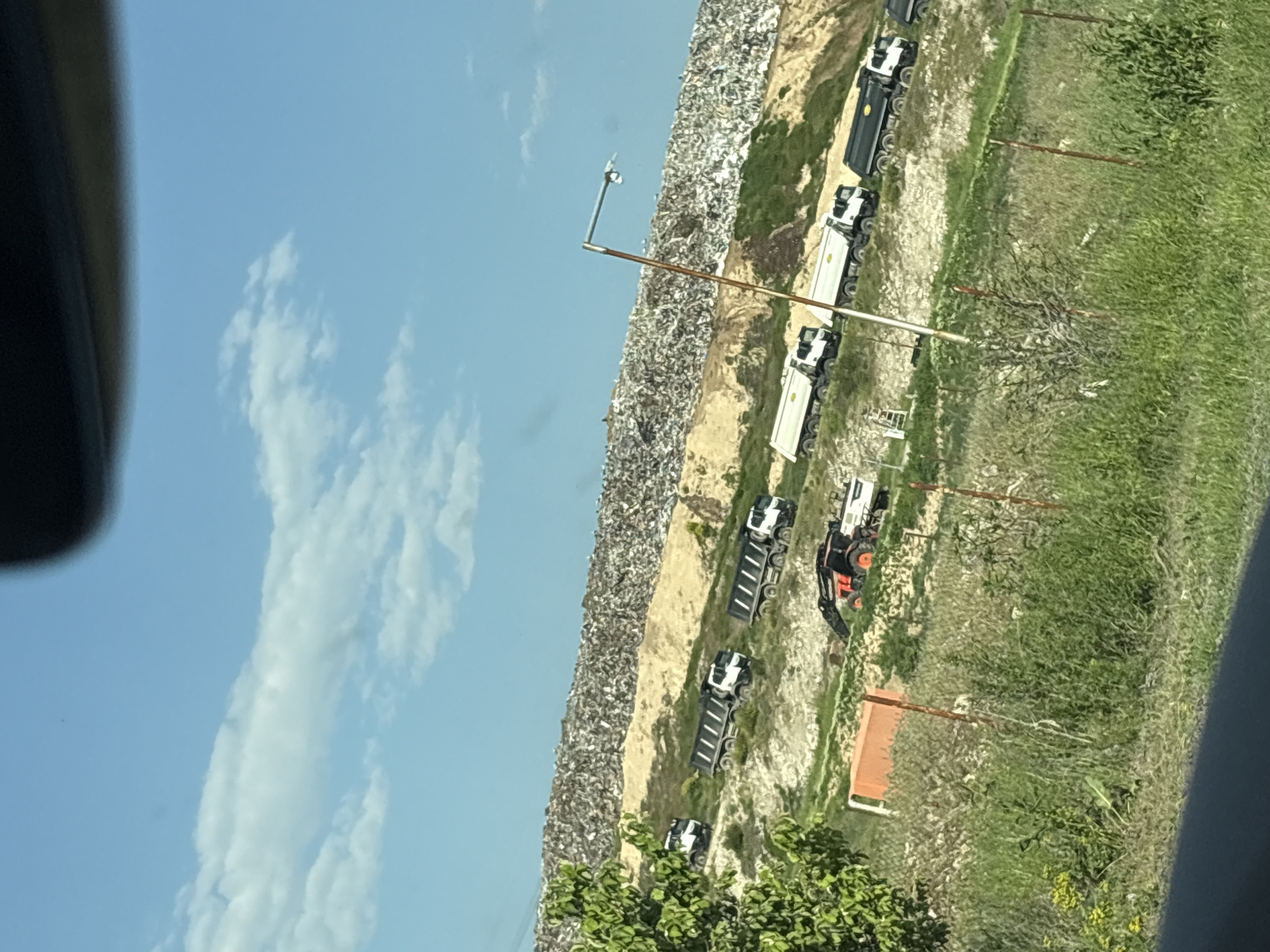The image depicts a landfill in Kapitan Dimitrievo, Bulgaria. A large pile of waste stretches along the left side of the frame. Several trucks are parked next to the landfill. To the right is a field with green grass and sporadic vegetation. A tractor is also visible. The sky is blue with some clouds. Parts of a vehicle are visible on both sides of the image frame, indicating that the image was taken from inside a car.