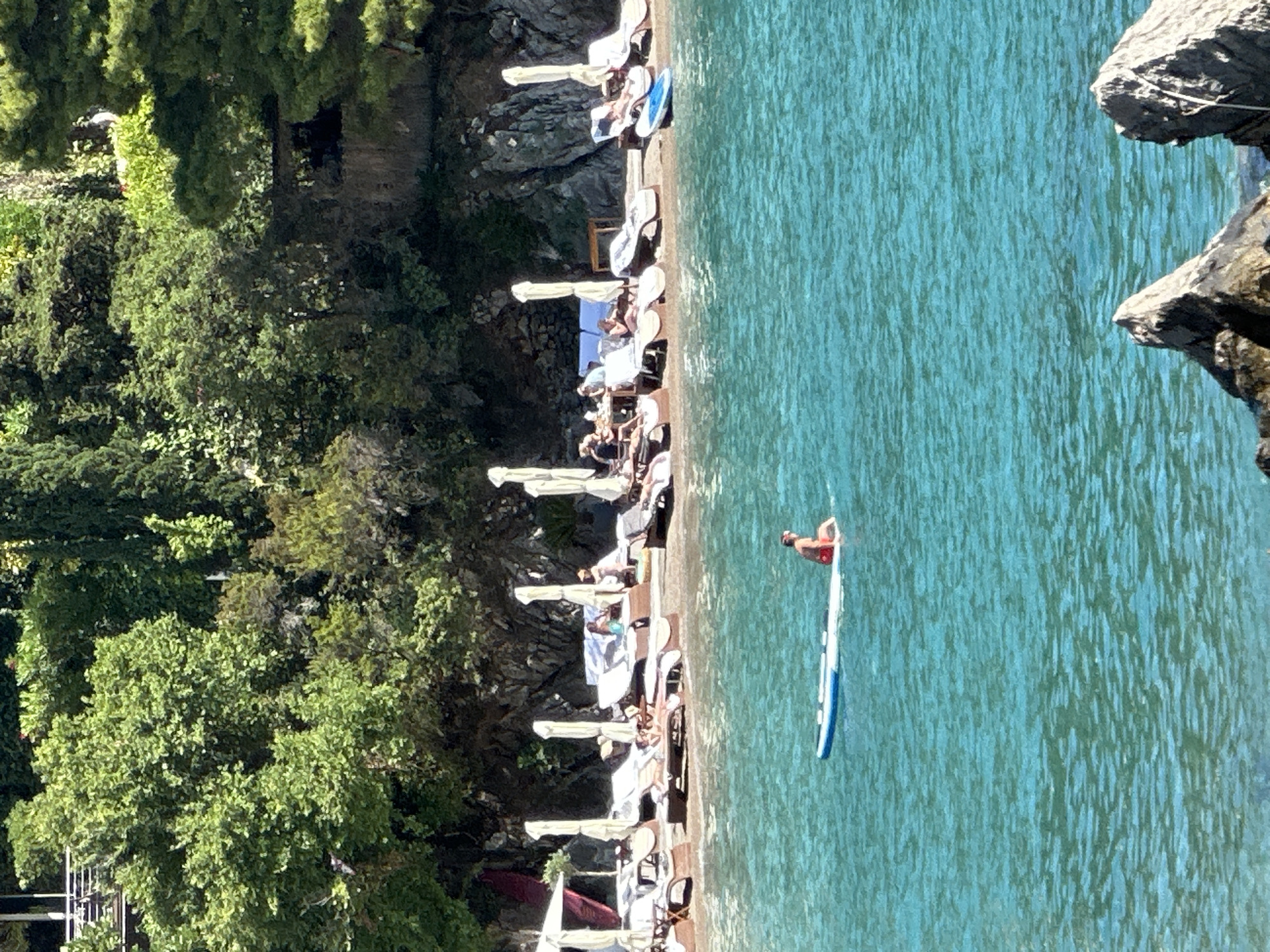 A man in red swim trunks is paddleboarding in the turquoise waters of the Adriatic Sea near Budva, Montenegro. In the background, a crowded beach is lined with lounge chairs and umbrellas. Lush green trees and rocky cliffs form the backdrop to the beach. The sun is shining brightly, creating a vibrant and inviting scene.