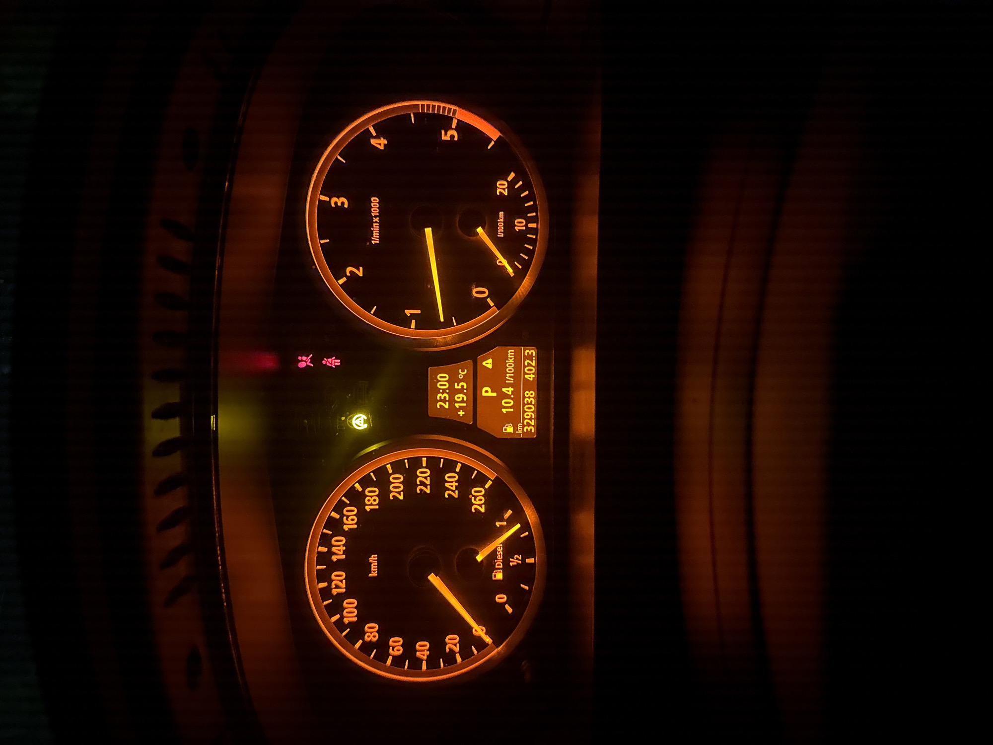 The image shows a car's dashboard at night. The speedometer reads approximately 20 km/h, and the tachometer is at 0 rpm. The fuel gauge indicates about half a tank of diesel. The display shows a time of 23:00 (11:00 PM), an outside temperature of 19.5°C, fuel consumption of 10.4 l/100km, and a mileage of 329038 km. It's in park (P). Specific warning lights are illuminated. There is no location information given, and thus I cannot determine the location in Stara Zagora, Bulgaria.