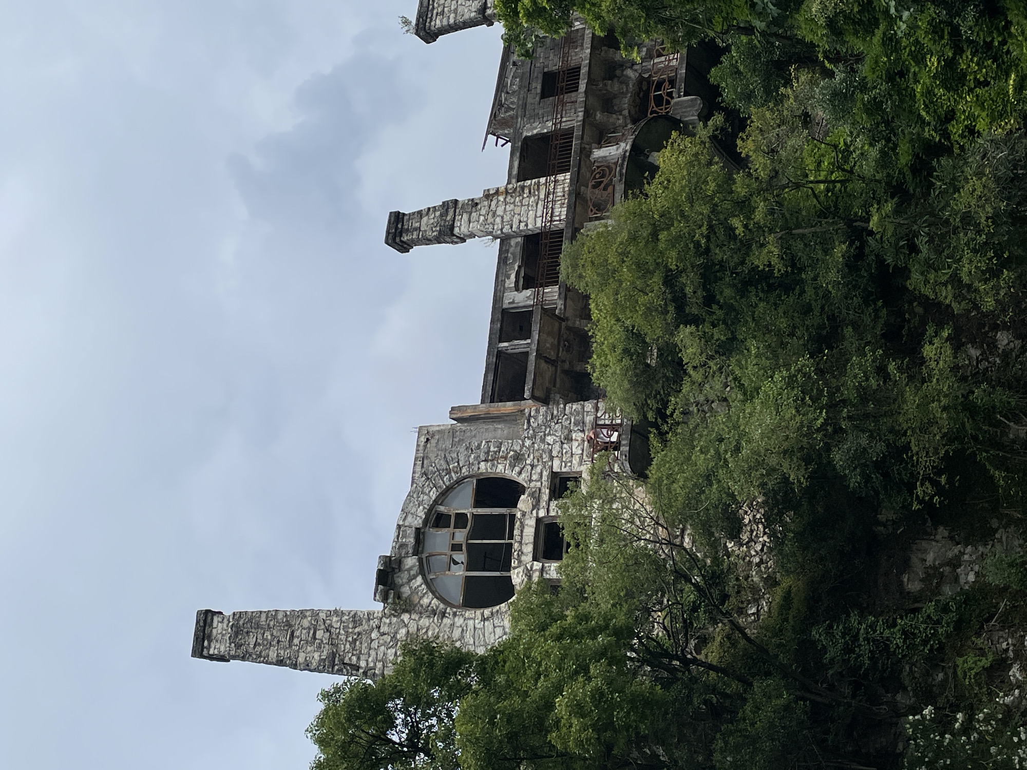 The image shows the facade of an abandoned, multi-story stone building, partially obscured by lush green trees and foliage. The building appears to be on a hillside. Distinct architectural features include a tall, rectangular stone chimney on the left, and a large, circular window with broken panes in the upper portion of the facade. Other elements visible are balconies with ornate railings, missing or broken windows, and crumbling stone work, indicative of decay. The sky above is overcast with a pale grey hue. In the middle ground, on a balcony, a person in a light-colored top and darker bottoms is visible. The building's setting suggests it is in a natural or semi-natural environment.