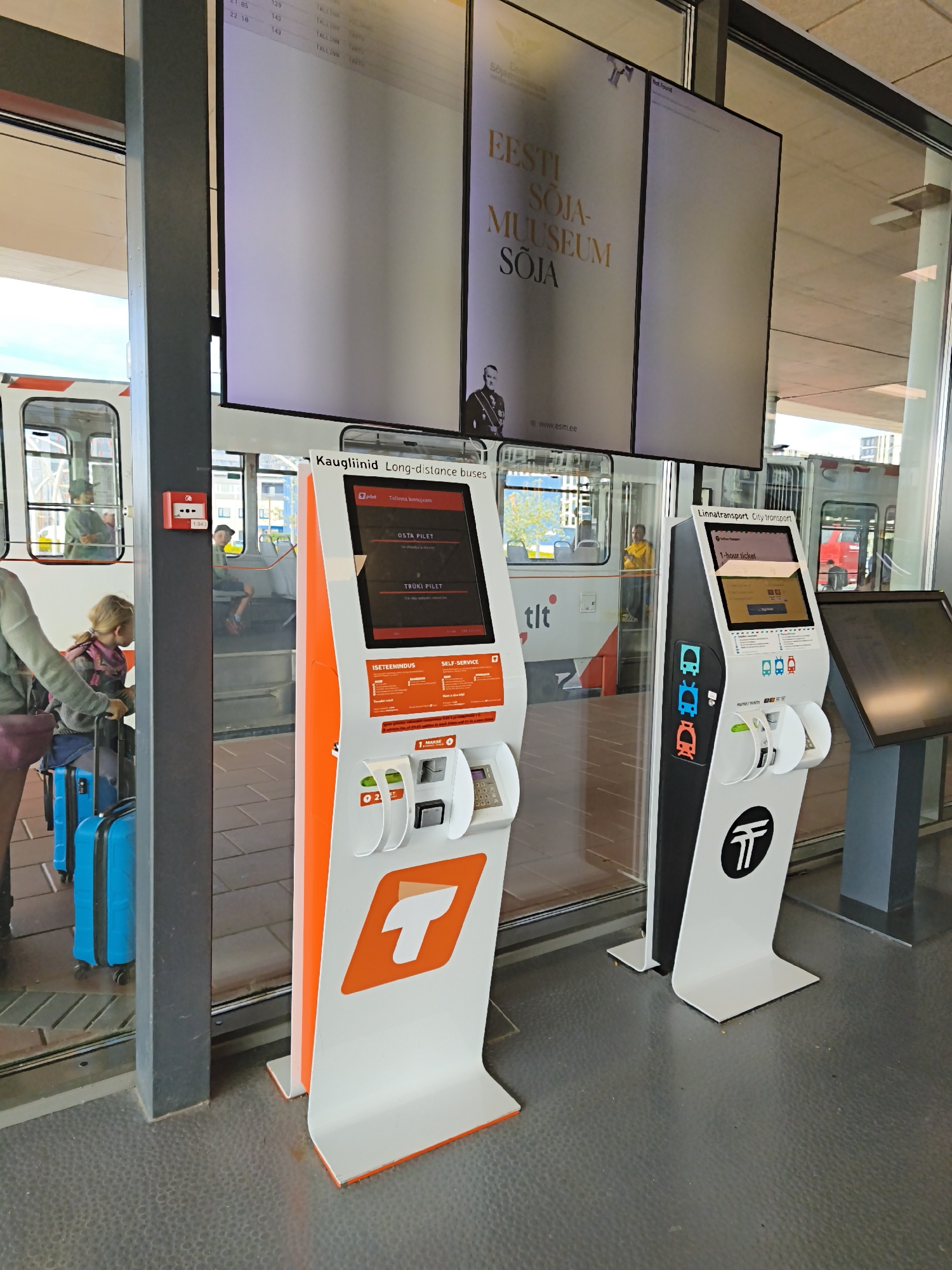 In Tallinn, Estonia, at a public transportation hub, two ticketing machines stand prominently. The machine on the left is orange and white, displaying options to "OSTA PILET" (Buy Ticket) and "TRÜKI PILET" (Print Ticket) in Estonian. Below these options, there are sections for "ISETTEENINDUS" (Self-Service) and payment. A young girl with blonde hair and a pink scarf is pulling two bright blue suitcases. Beside her, a person with a grey jacket stands, also near the ticketing machine.
To the right, a black and white ticketing machine advertises "Linna transport. City transport." and offers a "1-hour ticket." This machine features icons representing buses and trams and includes a section for "PUUTUMATA / TICKETS". It has a keypad for input. In the background, tram cars are visible through large windows, with passengers inside and on the platform. One tram has "tlt" branding. A large digital display screen hangs above, showing what appears to be departure information and an advertisement for the "EESTI SÕJA-MUUSEUM" (Estonian War Museum). The overall environment is modern and appears to be an indoor waiting area with grey tiled flooring.