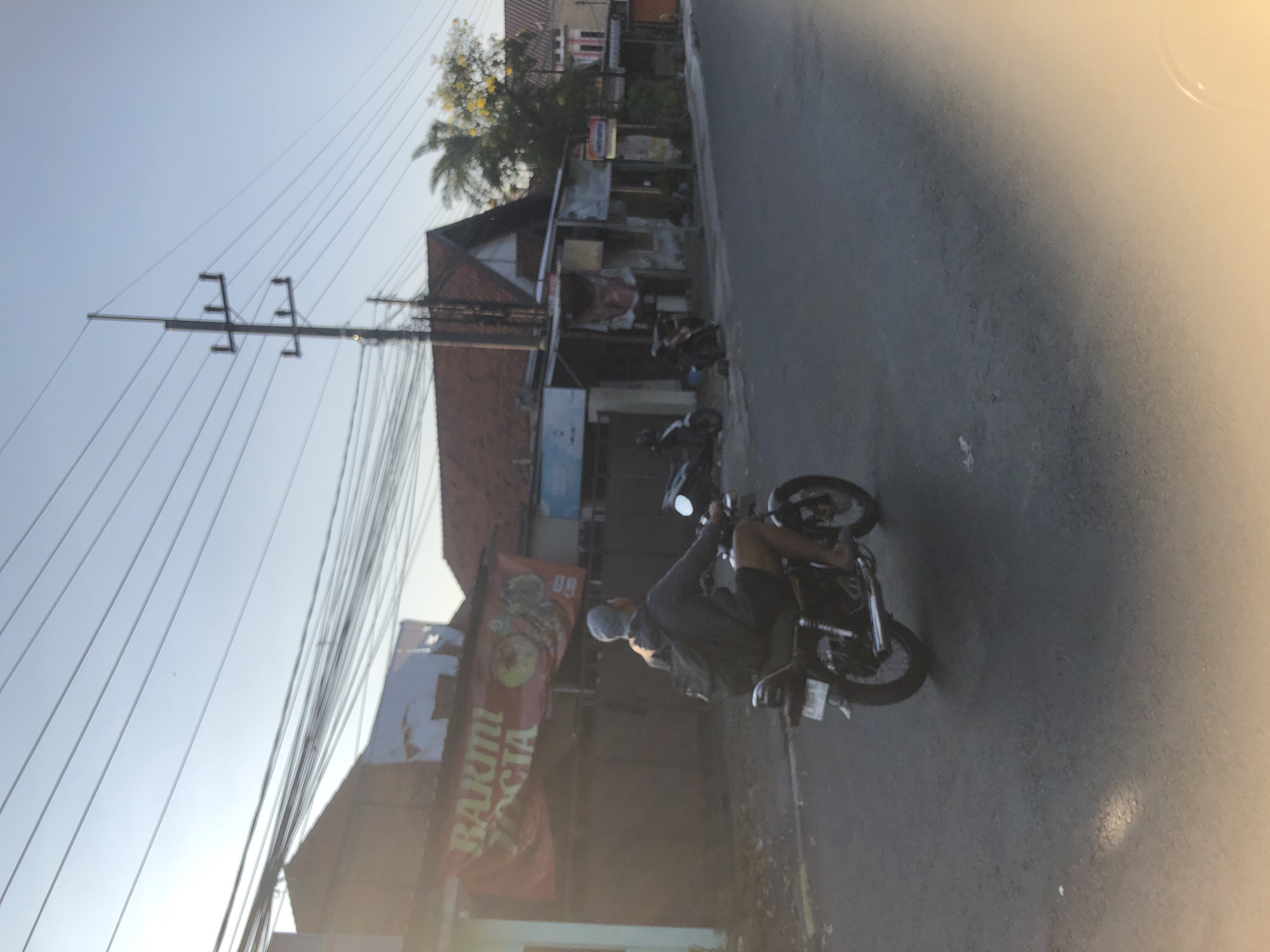 A person wearing a grey hoodie and a grey beanie is riding a dark-colored motorcycle down a paved road. The person is carrying a dark backpack. The motorcycle has a white license plate. In the background, there are buildings with red tile roofs, a banner that reads "BARTMI SOCIA", and utility poles with numerous wires. The sky is clear and blue, and the sun is low in the sky, casting long shadows. The scene appears to be in a residential or commercial area.
