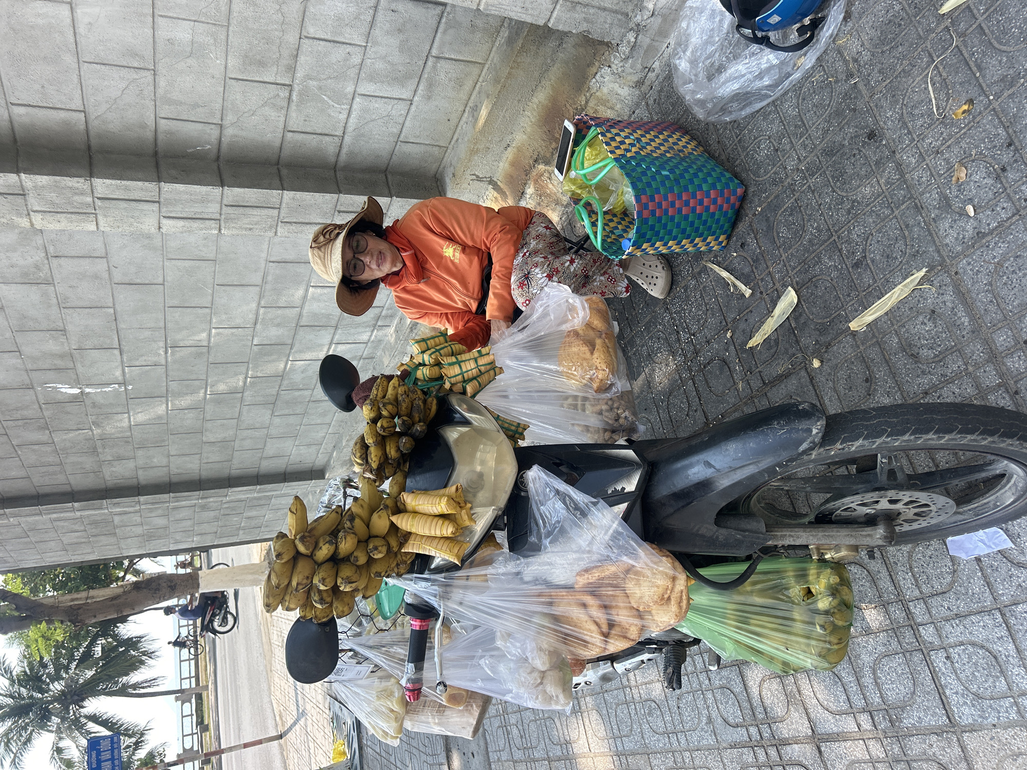 A woman wearing an orange jacket, floral pants, a hat, and glasses is squatting on a tiled sidewalk in Bắc Nha Trang, Vietnam. She is next to a black motorbike, which is laden with produce, including bunches of bananas, wrapped packages resembling rice cakes, and other unidentified items, all seemingly for sale. A woven basket sits near her, and a blue helmet is visible in the background. The background shows a palm tree and a glimpse of the beach across the street.