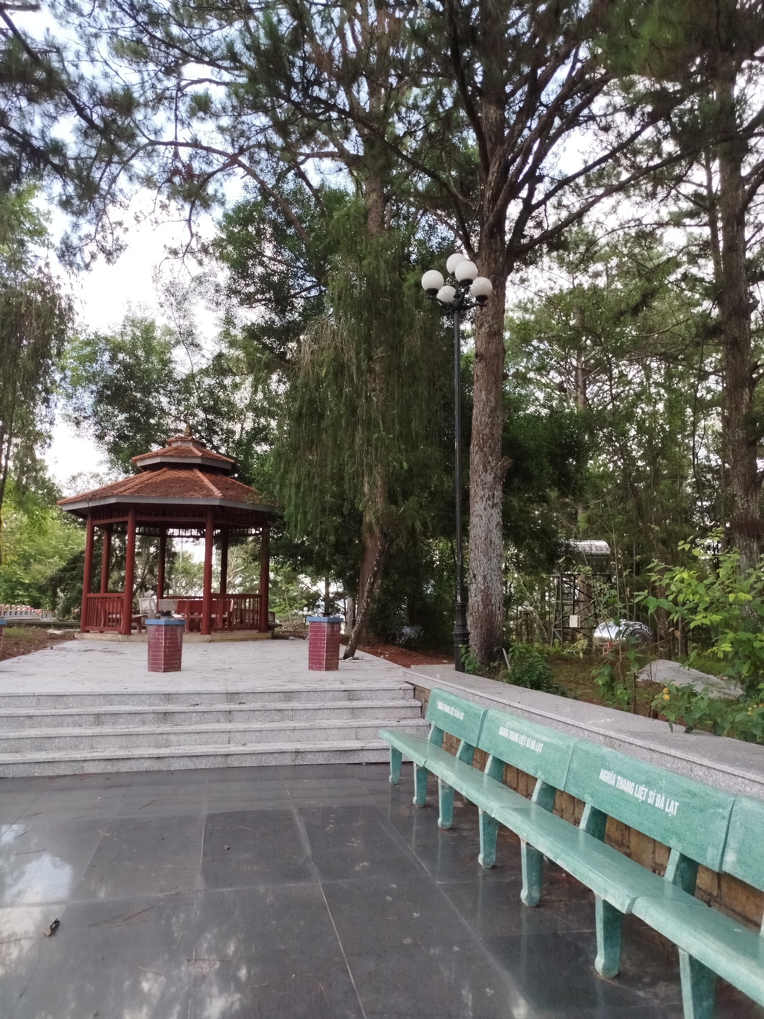The image depicts a park or memorial site in the city of Cam Ly - Đà Lạt, Vietnam. A red-roofed gazebo sits atop a raised platform accessible by steps. In the foreground are several green concrete benches. The area is surrounded by tall trees and vegetation. A streetlight stands near the trees. There are no people visible in the image. The ground is paved with dark gray tiles.
