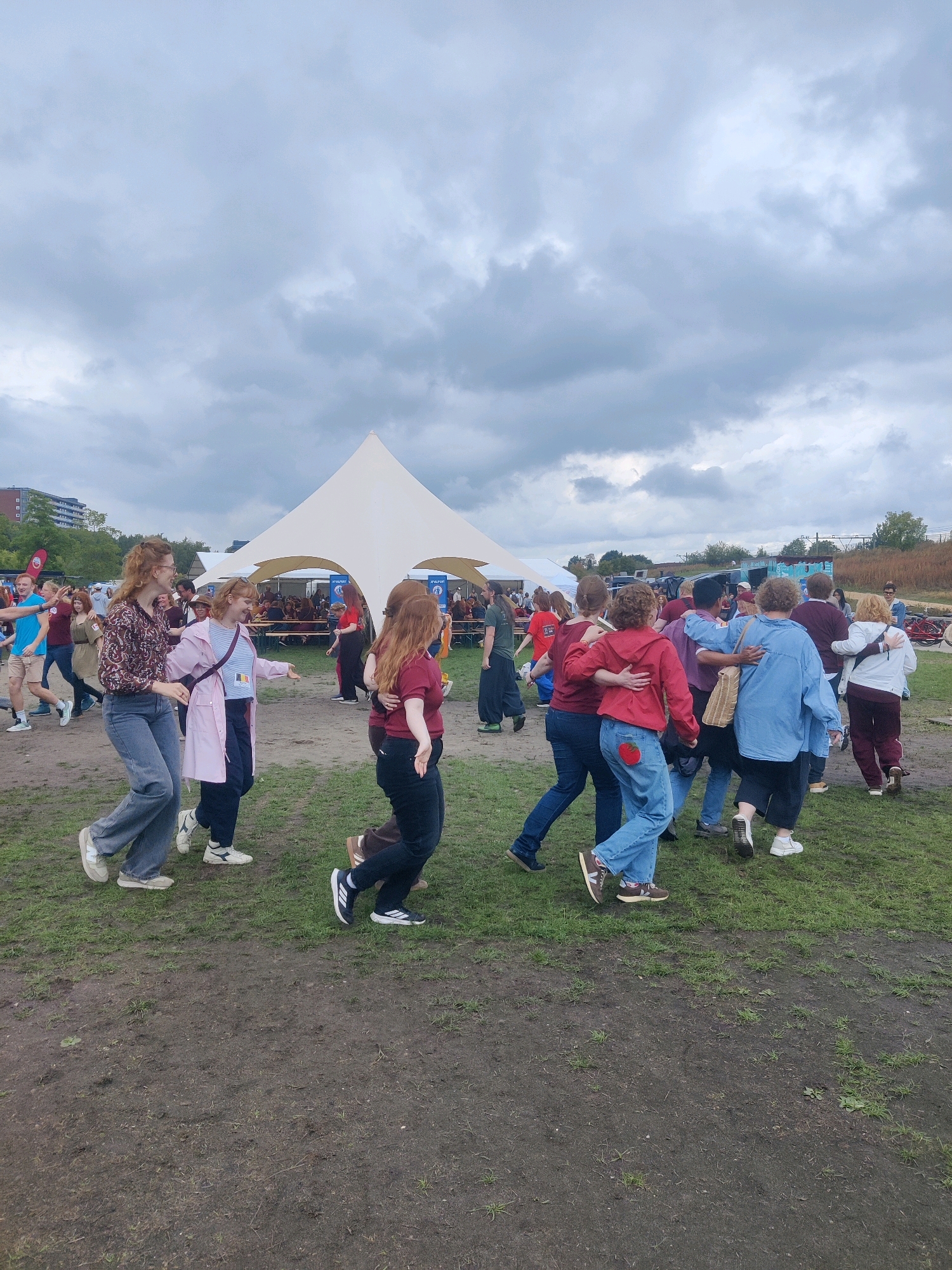The image depicts an outdoor event on a cloudy day. In the foreground, a group of people are dancing and interacting on a grassy and muddy area. They are casually dressed in jeans, t-shirts, and jackets. One woman on the left is wearing a patterned shirt and jeans, while a woman next to her is in a pink raincoat and jeans. Further to the right, a group is clustered together, with some individuals in red tops and blue jeans. One person is wearing a red jacket over a blue shirt. In the background, there is a large, white, conical tent structure, and beyond that, more tents, tables, and people sitting. There are also trees and buildings visible in the distance, suggesting an urban or semi-urban setting. The overall atmosphere appears lively and social, with a focus on the dancing group. The presence of a prominent white tent and the general ambiance could suggest this event is taking place at a festival or outdoor gathering in Tilburg, Netherlands, potentially near a park or open field.