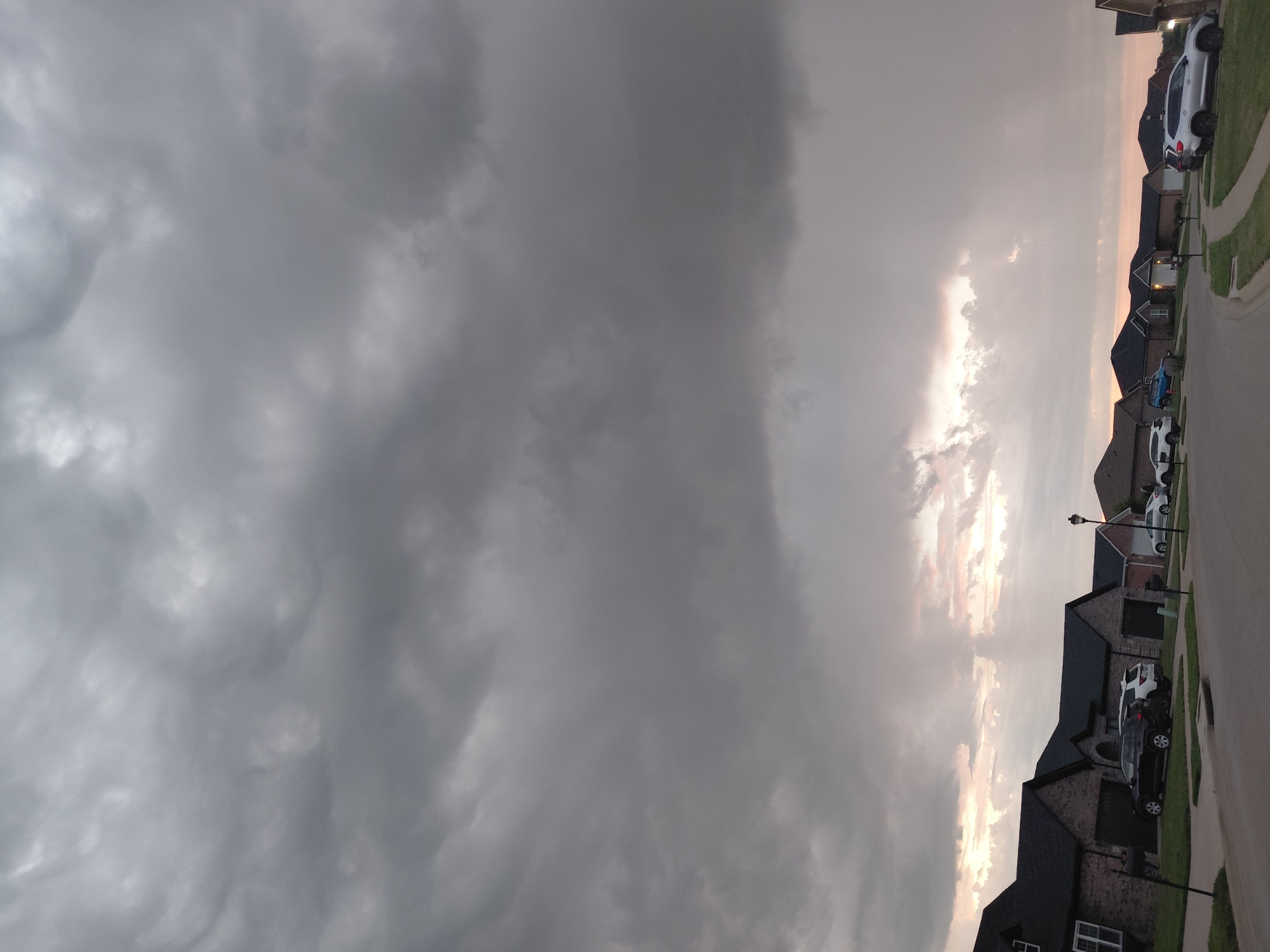 This is an image of a suburban street in Richmond, United States of America. A row of brick houses with dark roofs is visible on the left side of the street, with cars parked in the driveways. The street curves to the right. The sky is filled with dark, heavy clouds, suggesting an impending storm. The lower portion of the sky shows some lighter, brighter areas, possibly indicating the setting sun or a break in the clouds. No people are visible in the image.