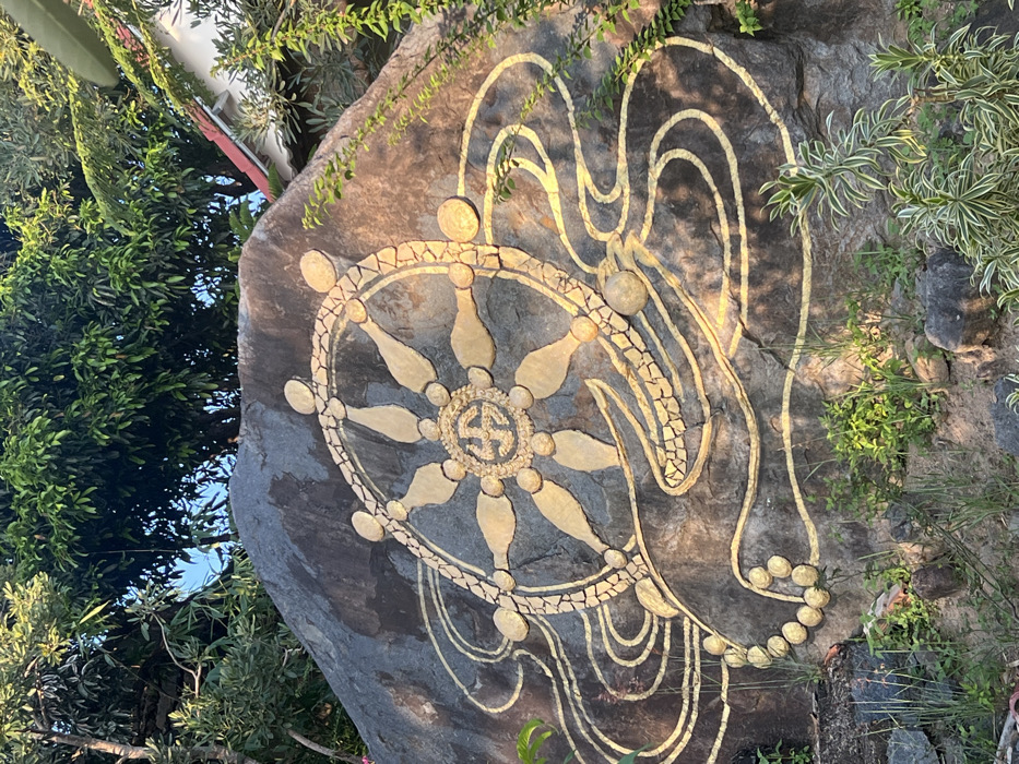 A large, dark grey rock, located in Nha Trang, Vietnam, is prominently featured, adorned with a gold-colored Buddhist Dharmachakra (Wheel of Dharma) emblem. The eight-spoked wheel is composed of gold-colored material, with its rim appearing to be a mosaic of small golden fragments. At the center of the wheel's hub is a gold-colored swastika symbol. Undulating, gold-colored lines, suggestive of water or clouds, emanate horizontally from beneath the wheel, extending across the rock's lower face. A string of spherical gold-colored elements is incorporated into these wavy lines on the lower left. The rock is set outdoors, surrounded by sparse green ground vegetation, grass, and exposed soil at its base. Above the rock, dense green tree foliage is visible. To the upper right, partially obscured by leaves, a structure with a red tiled roof and white eaves is visible. Direct sunlight illuminates the golden artwork, casting subtle shadows on the rock's surface. No individuals or specific actions are depicted.