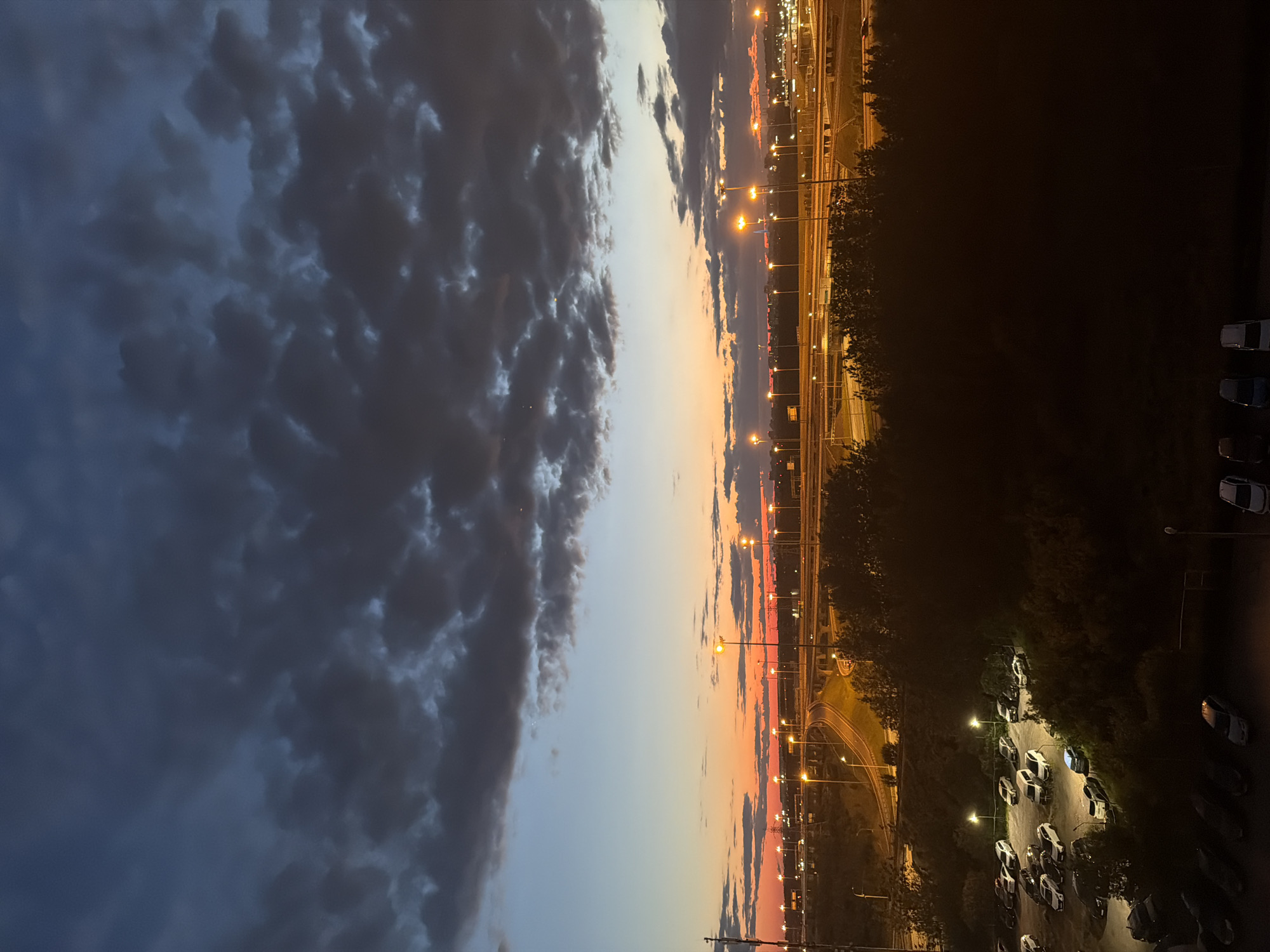 The image depicts an aerial view of a highway interchange and a parking lot at dusk in the city of Unknown, Unknown. The sky is a gradient of colors, transitioning from a deep blue at the top to orange and red near the horizon, with dark, textured clouds scattered throughout. Numerous streetlights illuminate the highway and roads, casting a warm glow on the asphalt and surrounding landscape. The highway appears to be a multi-level structure with several on and off ramps. Below the highway, a dark, dense line of trees forms a natural barrier. In the foreground, a dimly lit parking lot is visible, with several cars parked in designated spots. The overall scene suggests a transition from day to night in an urban environment.