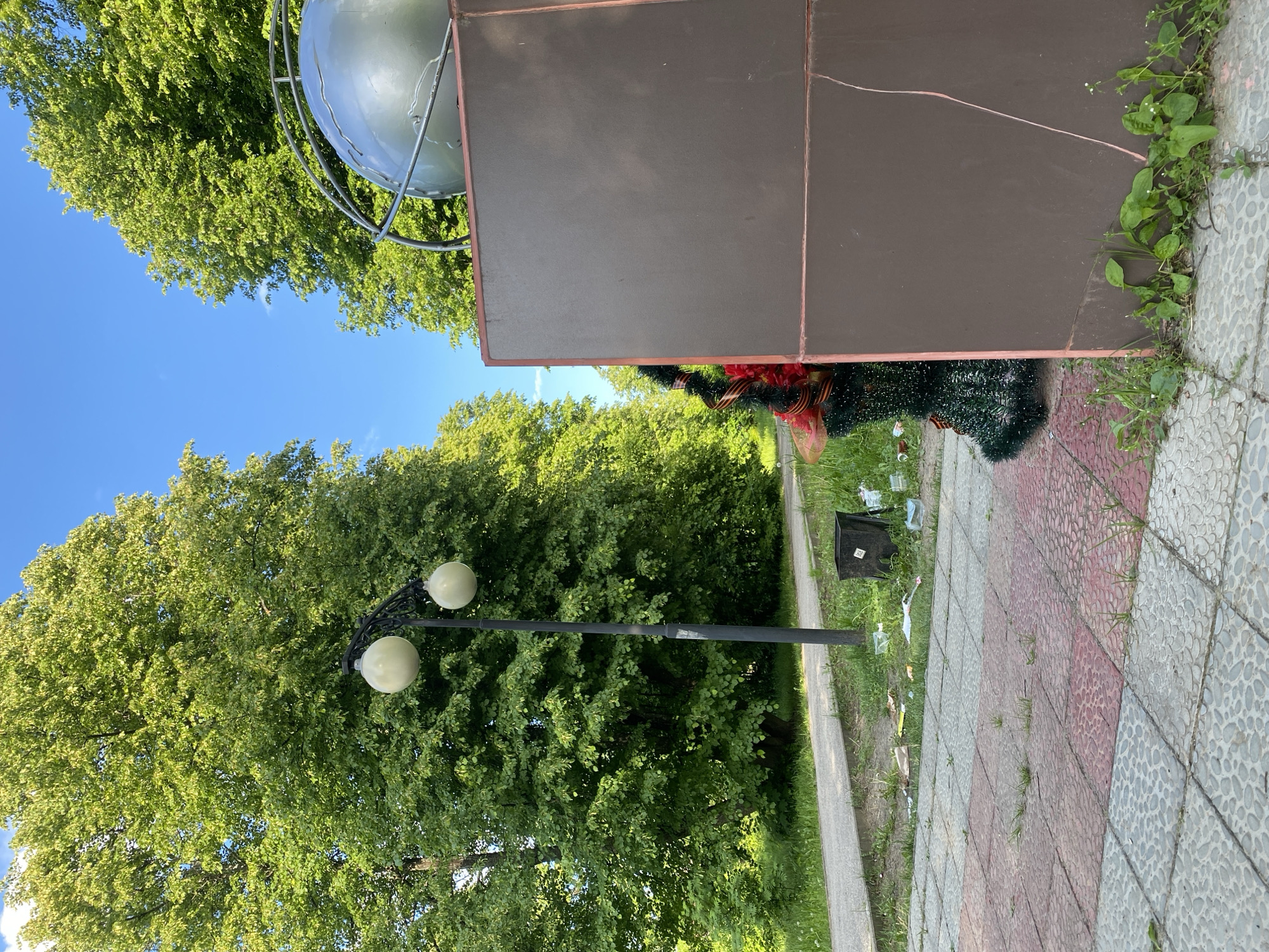 The image captures an outdoor public space on a clear, sunny day. A vast canopy of bright green leaves from numerous trees fills the background and much of the left side of the frame, contrasting with a vibrant blue sky visible in the upper left corner.
In the mid-ground, a dark, metal street light featuring two spherical white lamps stands upright. To its right, a large, dark rectangular monument base, possibly made of panels, occupies a significant portion of the center-right. This base has reddish-brown trim along its edges. Partially visible above this base is a silvery metal globe sculpture, framed by a metal support structure, reflecting the sunlight.
At the base of the monument, on the right side, a dark green decorative garland adorned with red accents is visible. The foreground features a paved area constructed from various square and rectangular tiles in shades of red, gray, and speckled patterns. A narrow, lighter concrete path runs parallel to this paved area on the left.
Near the concrete path, a black waste bin is present, surrounded by visible litter including several plastic bottles and other debris. Small green plants and weeds are growing in the cracks of the pavement and along the base of the monument, suggesting a lack of maintenance in parts of the area. No people are visible, and there is no discernible text.