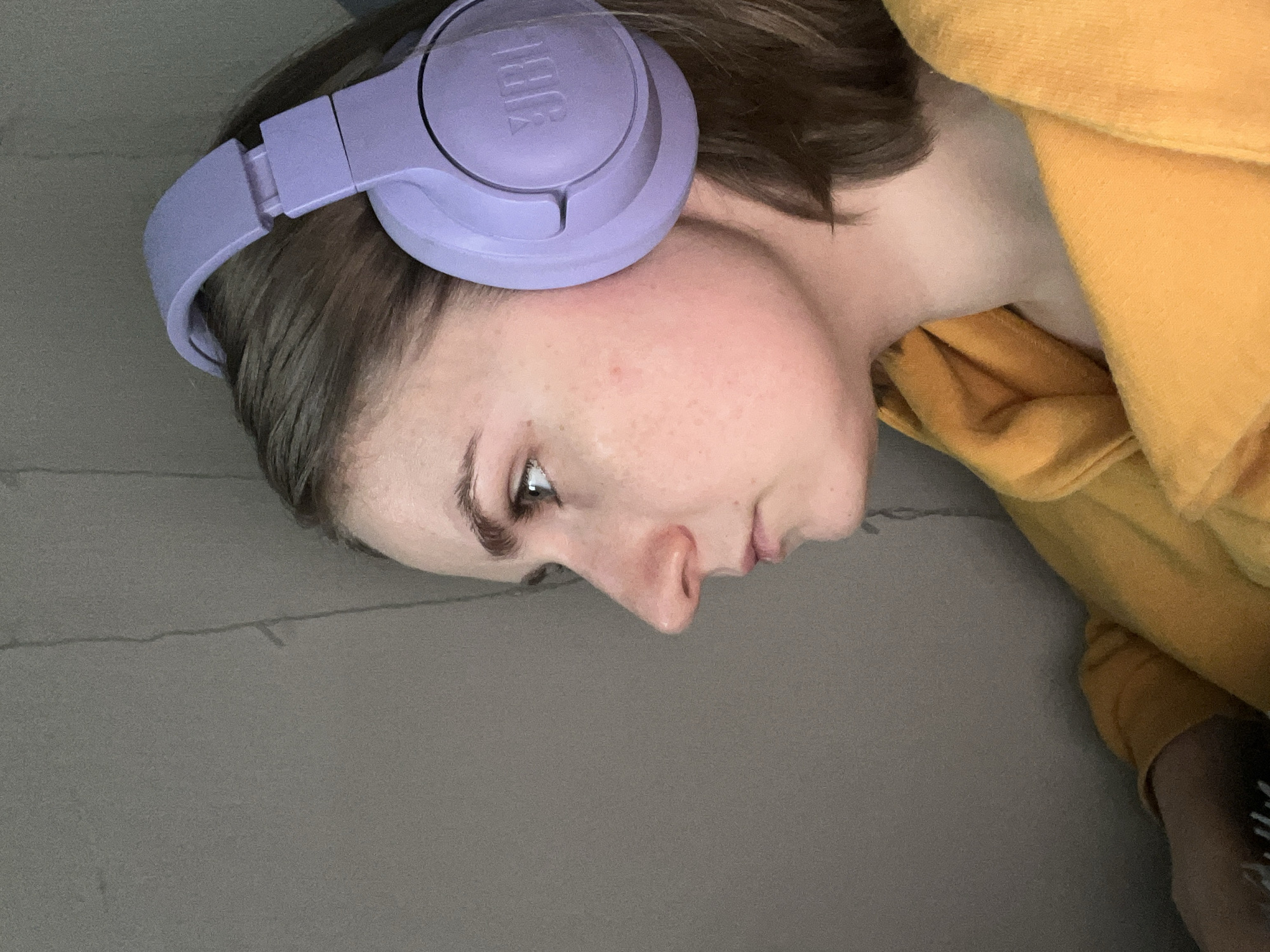 A close-up image shows a person lying on a gray surface, possibly a bed or couch. The person is wearing purple JBL headphones and a yellow hoodie. The location is not discernible from the image.