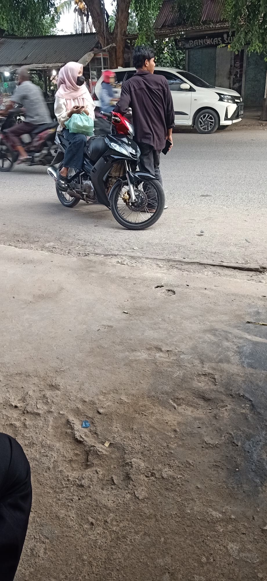 In Gampongteungoh, Indonesia, a young woman wearing a light pink hijab and a black face mask is seated on a black motorcycle. She is holding a light green plastic bag in her lap and has her right hand on her phone. Beside her, a young man in a dark long-sleeved shirt and dark pants stands, holding his phone in his right hand. The motorcycle has red accents on the front and silver spokes on the wheels. In the background, a white car is parked, and a building with a sign that appears to read "Pangkas Su" is visible. Trees with lush green foliage line the street, and another person on a motorcycle is blurred in motion to the left. The foreground shows a rough, textured concrete surface with some debris and tire tracks. The scene appears to be a roadside setting, possibly near a shop or business.