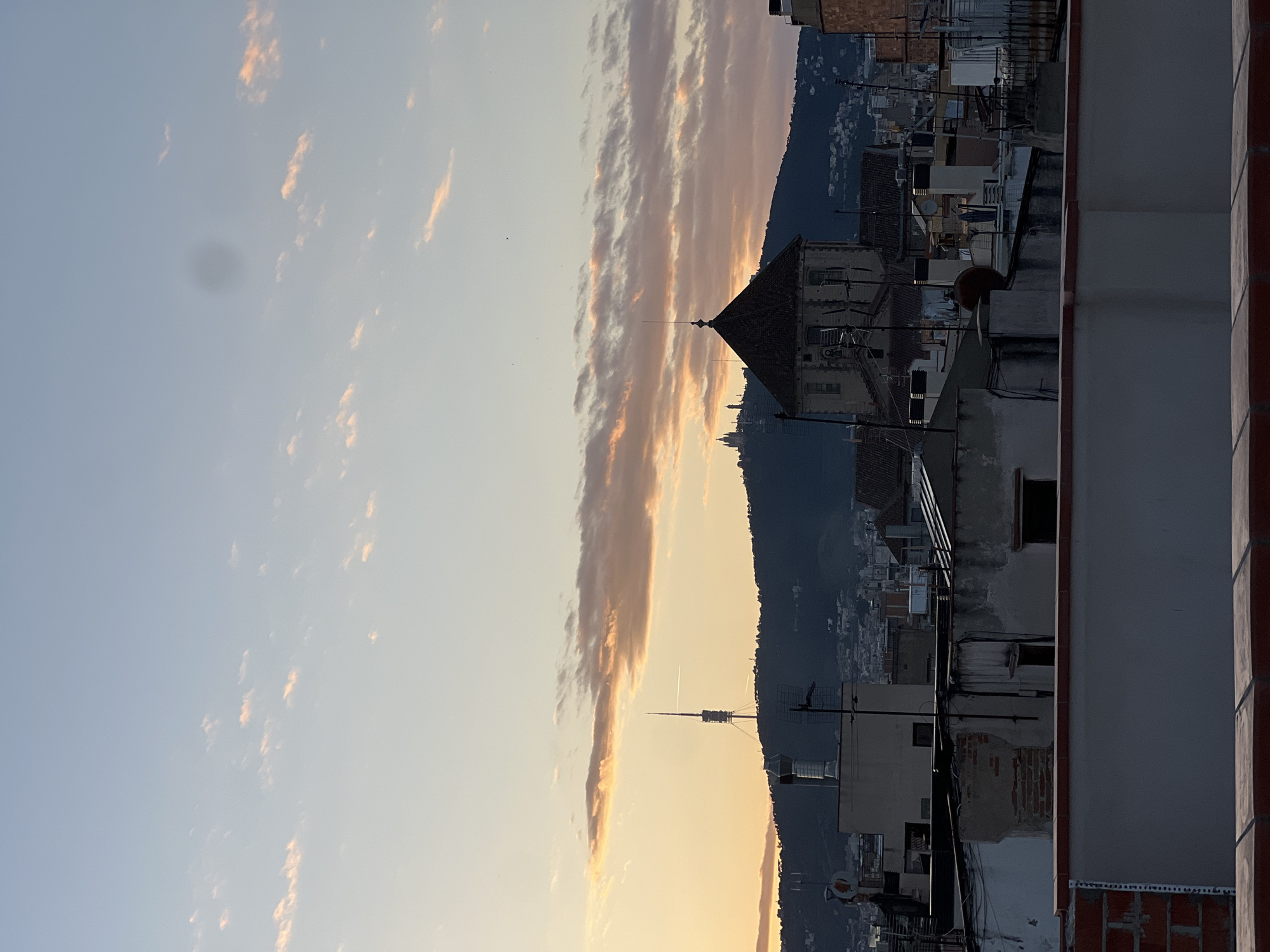 The image captures a view from a rooftop in Barcelona, Spain, at dusk. A long, dense cloud formation runs parallel to a mountain range, bathed in the orange light of the setting sun. The cityscape is visible in the foreground with various buildings and rooftops in varying states of repair. An antenna tower stands against the backdrop of the mountain. The overall atmosphere is urban, quiet, and tinged with the colors of sunset. The image is slightly out of focus, especially in the sky. There are no people visible in the shot.