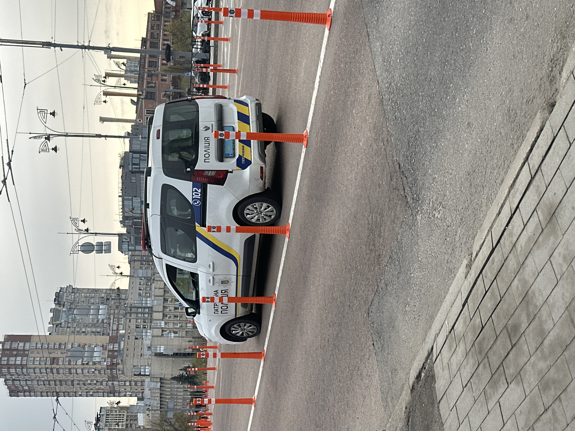 The image displays an urban scene in Kyiv, Ukraine, centered around a white Ukrainian Patrol Police van. The vehicle, likely a Renault, is situated on an asphalt surface, possibly a training ground or a cordoned-off area, and is bordered by a continuous white painted line. Numerous orange and white traffic cones are strategically placed around and in front of the police vehicle, suggesting a driving course, temporary barrier, or designated parking.
Visible text on the police van includes "ПАТРУЛЬНА ПОЛІЦІЯ" (Patrol Police) on the side, "ПОЛІЦІЯ" (Police) on the rear, and the emergency number "102." The Ukrainian coat of arms (tryzub) is also prominently displayed on the side of the vehicle.
In the background, a dense urban landscape features multiple multi-story residential or commercial buildings under an overcast or cloudy sky. Overhead wires, indicative of a public transport system like trolleybuses or trams, and decorative streetlights are visible, spanning across the scene. To the right, a paved sidewalk with rectangular tiles borders the asphalt road.
The scene takes place during daytime, characterized by soft, diffused lighting consistent with cloudy weather. The overall impression is one of a functional urban space, possibly used for police training or operations.