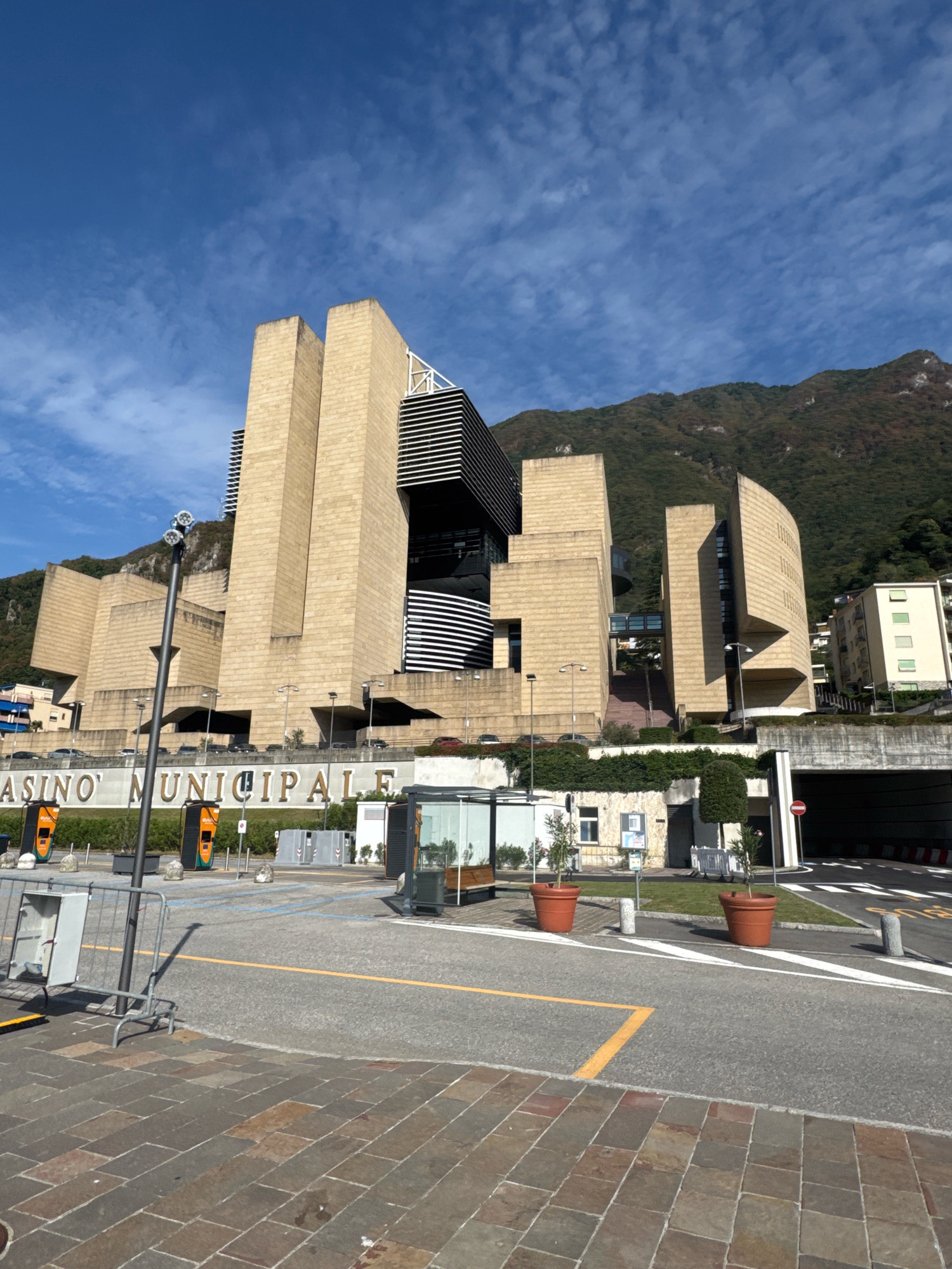 This is a wide, low-angle, exterior, eye-level shot of the Casino Municipale in Campione d'Italia, Italy, on a clear, sunny day with a few wispy clouds in a bright blue sky. The casino is a large, modern building with a distinctive brutalist architectural style, constructed from pale, textured stone blocks. The building features geometric shapes, with several tall, rectangular towers and a central section with horizontal black louvers. A curved, lighter-colored section is visible on the right side of the building. The name "CASINO MUNICIPALE" is prominently displayed in large, light-colored letters on a wall in front of the building.
In the foreground, there is a paved area with a brick-like pattern, leading to a grey asphalt surface with yellow and white road markings. Several streetlights with multiple bulbs are present. Two large, orange electric vehicle charging stations are visible on the left side of the paved area. Metal barriers and some potted plants in large, terracotta-colored pots are also in the foreground. In the middle ground, there's a covered bus stop shelter and some greenery. A tunnel entrance is visible on the right side of the image, with a red and white striped barrier. In the background, the casino building dominates the scene, with a steep, tree-covered mountain rising behind it. To the right of the casino, a multi-story residential building with light-colored walls and green-framed windows is visible. The overall environment suggests a modern urban setting integrated with a natural mountainous landscape. There are no people visible in the scene.