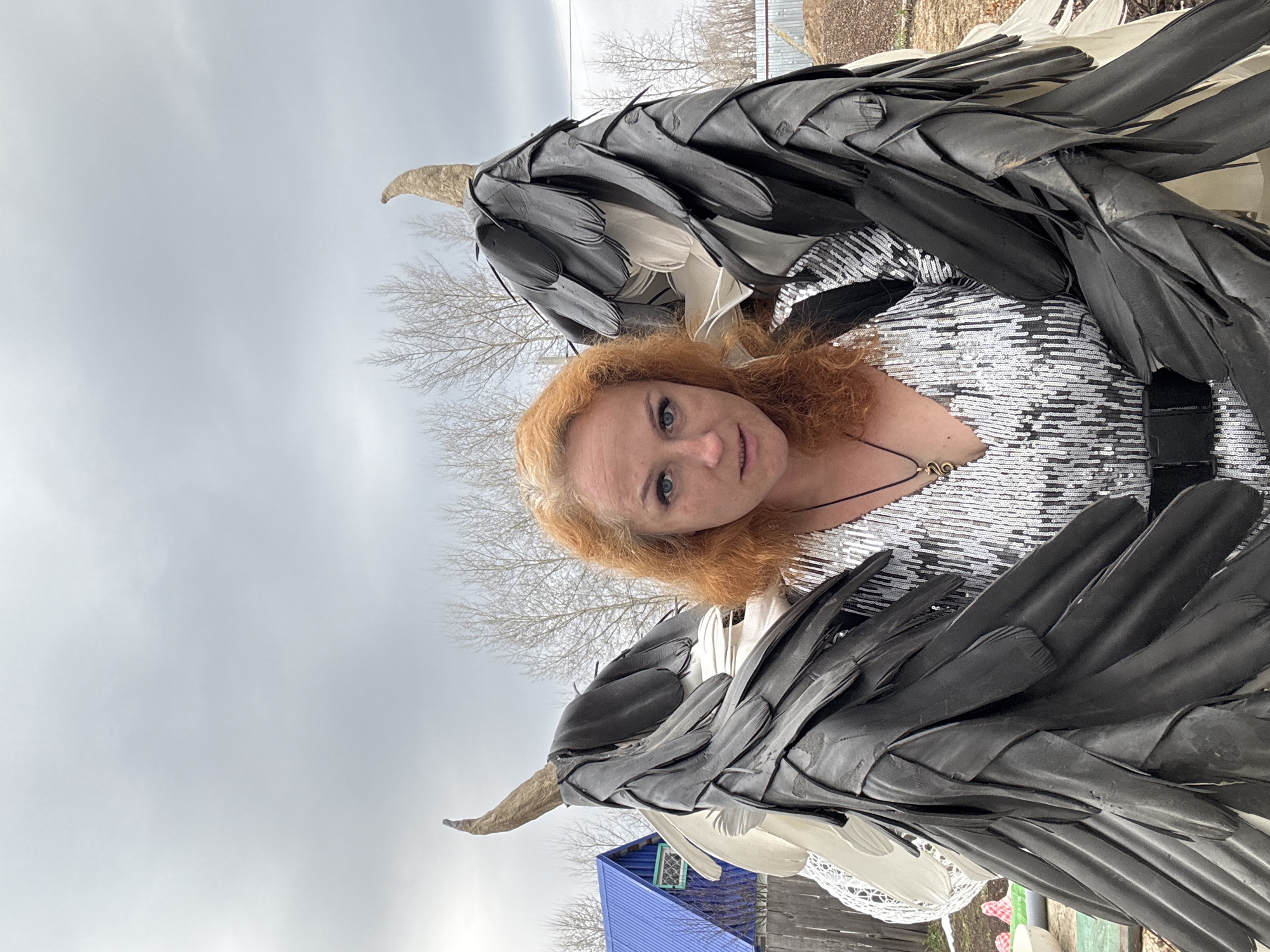A woman with red hair and blue eyes poses for a close-up shot in Chaikovsky, Russia. She wears a silver sequined top. Large, dark, textured wings are attached behind her, partially obscuring the background. The sky is overcast. In the background are bare trees, a wooden fence, and a blue structure. The image appears to be taken at a low angle.
