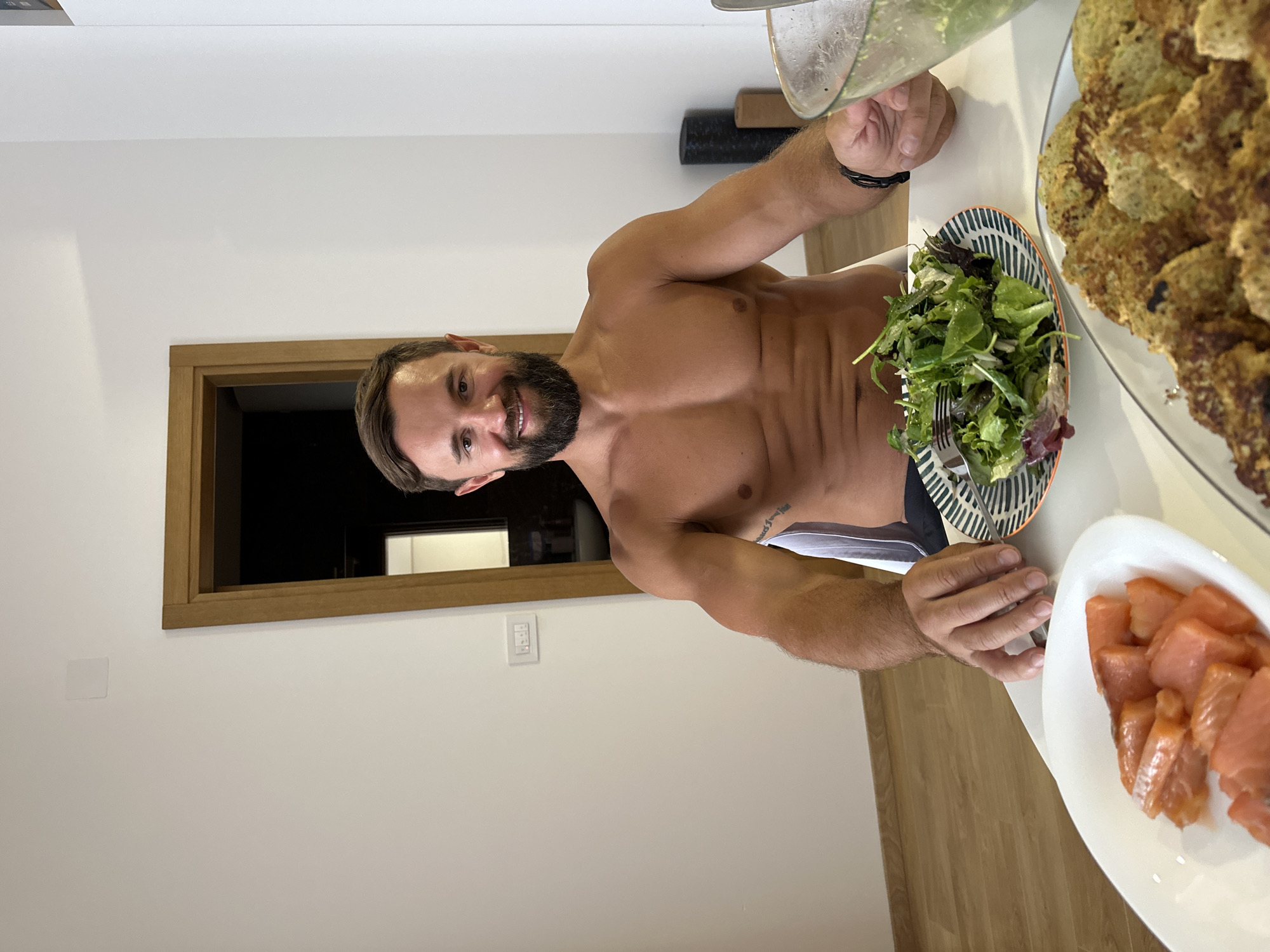 In this image, a shirtless man with a beard is seated at a table, preparing to eat. He is wearing black underwear. In front of him are two plates: one with a salad and a fork, and another with several slices of smoked salmon. To his right, a plate is filled with what appear to be fritters or pancakes. The man is smiling, and his demeanor suggests he is enjoying himself. The setting appears to be a modern, well-lit room, possibly a dining area or kitchen, with a wooden door frame visible in the background. The overall atmosphere is casual and relaxed. The time of day is not explicitly indicated, but the indoor lighting suggests it could be evening or a dimly lit daytime. There are no visible signs or text in the image.