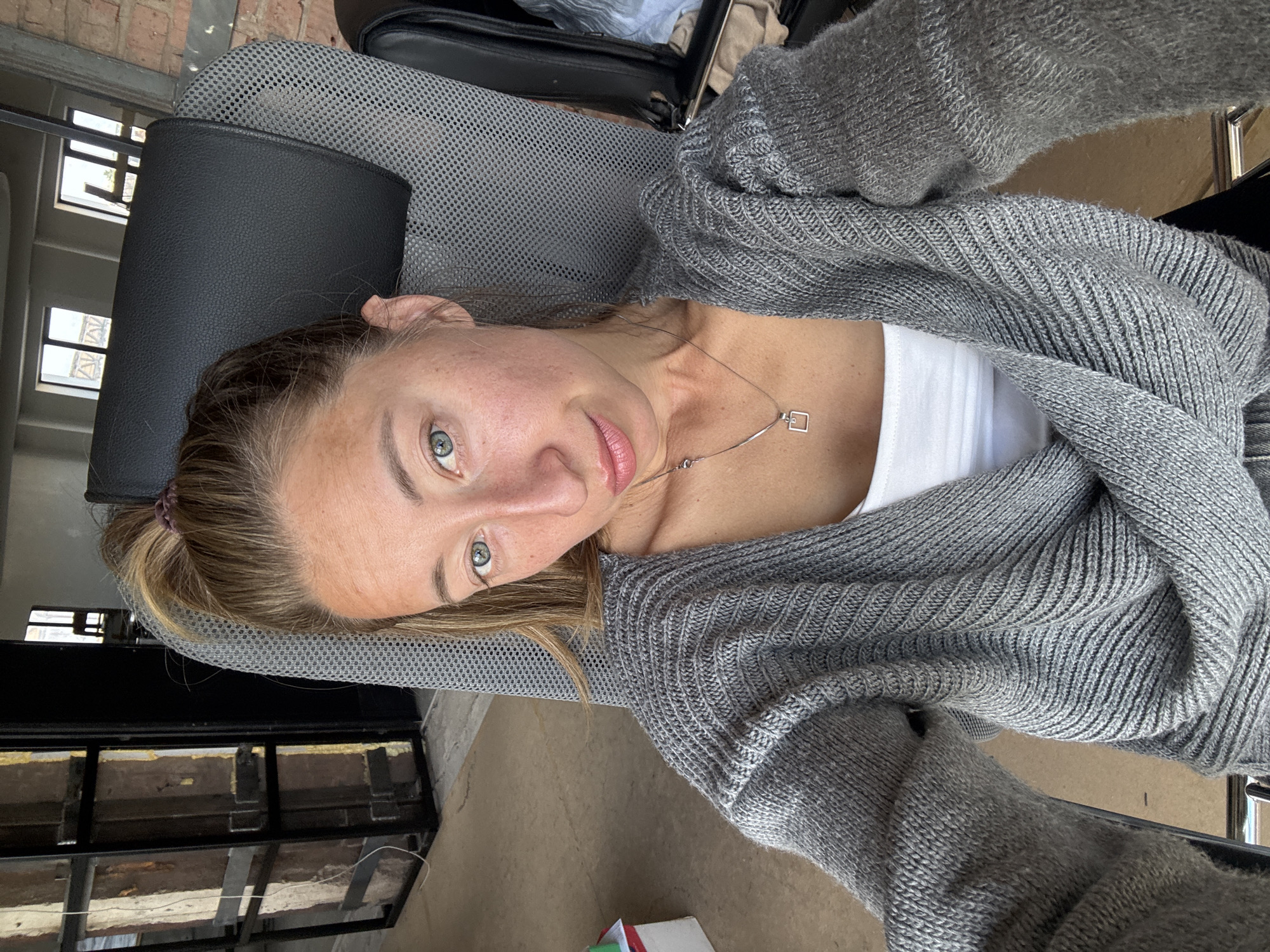 A woman with light brown hair pulled into a ponytail is seated in a gray office chair, facing the camera. She is wearing a gray knitted cardigan over a white tank top, and a silver necklace with a square pendant. She has light skin, green eyes, and light pink lips. The background shows an indoor industrial-style space with exposed brick walls and large windows. To the left, there is a tall metal shelving unit with several compartments. The floor appears to be concrete. The scene suggests an office or workshop environment in Chaikovsky, Russia.