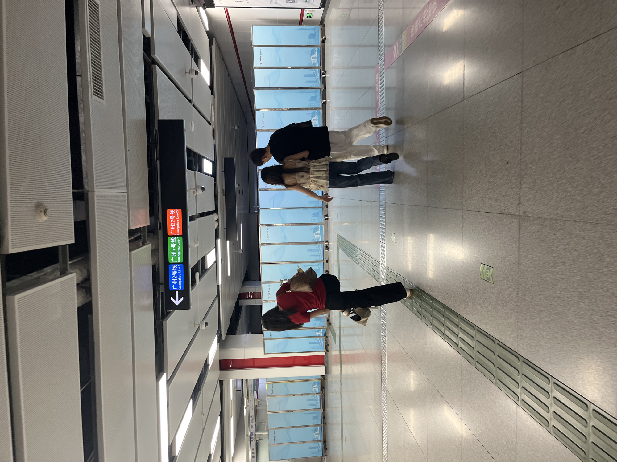 The image depicts a modern subway station in Panyu District, China. In the foreground, a woman in a red shirt and black pants is walking away from the camera, carrying a bag. Behind her, a couple, a man and a woman, are walking in the same direction, with the man's arm around the woman's shoulder. They are walking on a light grey tiled floor with tactile paving strips. In the background, there are several tall, light blue folding screens with white graphic designs, and a sign above indicates directions to "Guangzhou Line 2", "Guangzhou Line 7", and "Guangzhou Line 22". The station has a high ceiling with white perforated panels and fluorescent lighting. The overall environment is clean and well-lit.