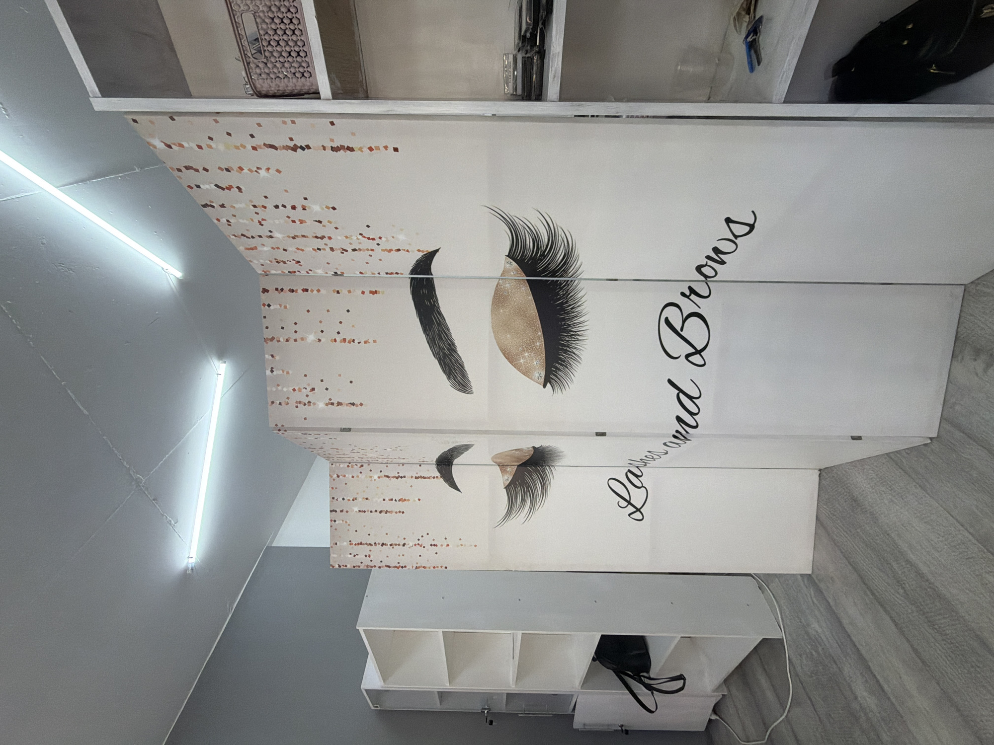 The image shows a room with white shelving units, a decorative screen with images of eyes and eyelashes, and a light fixture on the ceiling. The screen is partially visible, showing the words "Lashes and Brows" in a script font. A black bag is on the bottom shelf of the shelving unit on the left. The floor is covered with grey wood-look laminate flooring. The walls appear to be painted in a light grey and white. The lighting suggests an indoor commercial space, possibly a beauty salon. The overall impression is of a clean and organized setting.