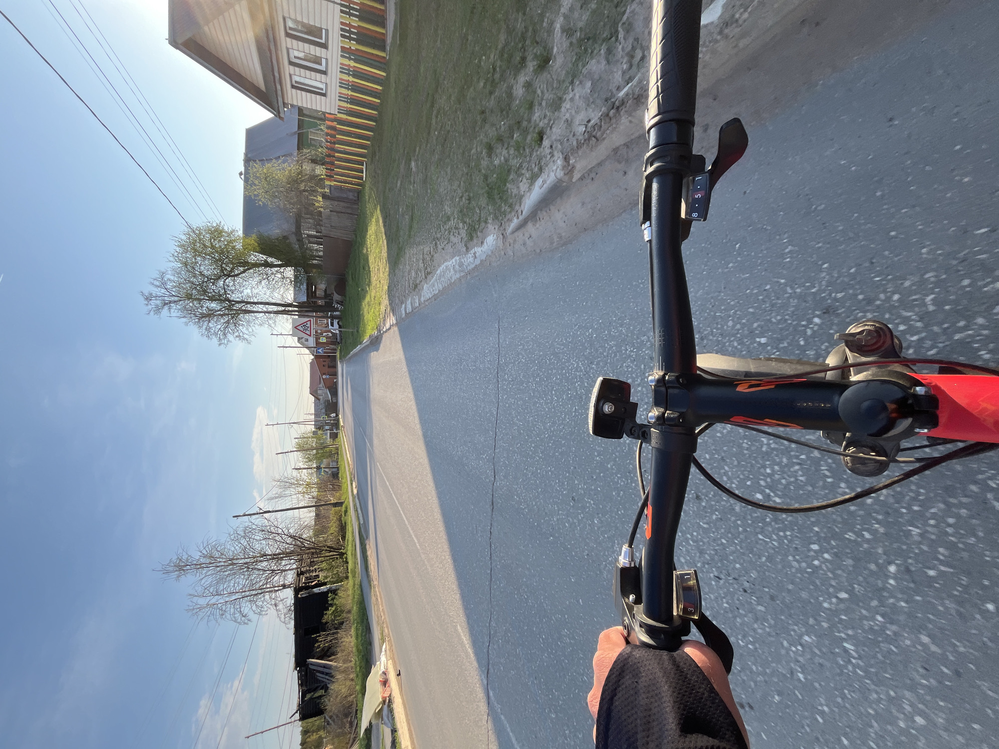 A first-person perspective image captures a bicycle ride on an asphalt road in the city of Chaikovsky, Russia. The handlebars of the bicycle are prominently visible, featuring gear shifters, a black-cased light, and a hand gripping the handle. The road stretches forward, marked by a significant crack running along its surface. To the right of the road, there is a grassy verge. On the left, a wooden fence, painted with vibrant vertical stripes, separates the road from a grassy area containing a house. In the background, trees line the road, extending the perspective. Overhead, power lines stretch across the clear, blue sky.