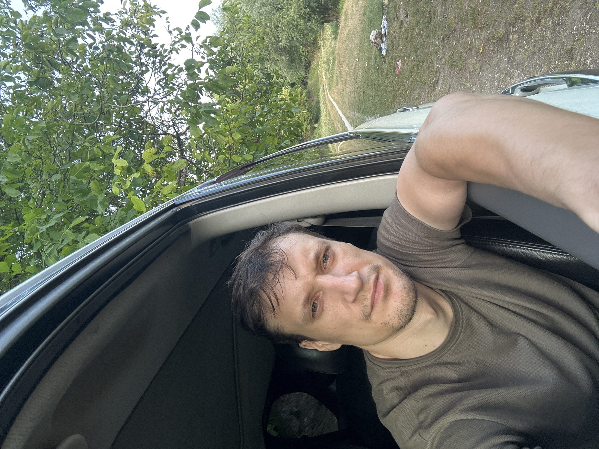 A man with brown hair and a light complexion is looking out of a car window. He has a serious expression on his face and is wearing a dark olive-green t-shirt. His left arm is resting on the open window frame. The car appears to be parked on a dirt road next to a grassy area with trees. There are some items of trash scattered on the ground to the right of the car. The background shows a dense canopy of green leaves from trees. The location is specified as Makiv, Ukraine.