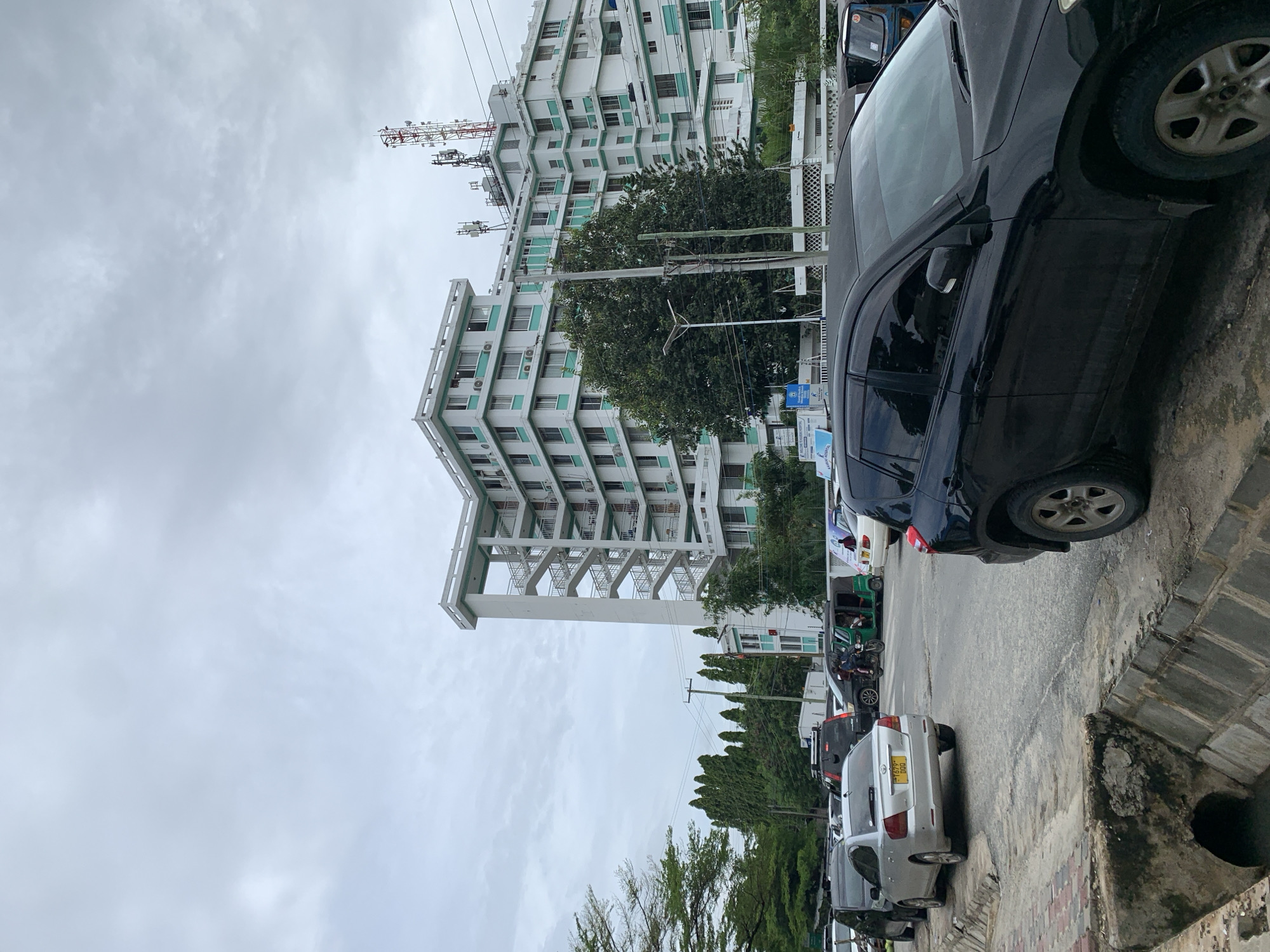 The image shows a street scene in Dar es-Salaam, Tanzania. The view is angled upwards to capture a tall, multi-story building with a white facade and green window frames. A network of communications towers is visible atop the building. Along the side of the street, several cars of various makes and models are parked, including a black sedan and a silver hatchback with a Tanzanian license plate. The street itself appears to be paved, with a concrete curb and drainage system. Trees and utility poles line the street, and the sky is overcast with a gray, cloudy sky. No people are clearly visible.
