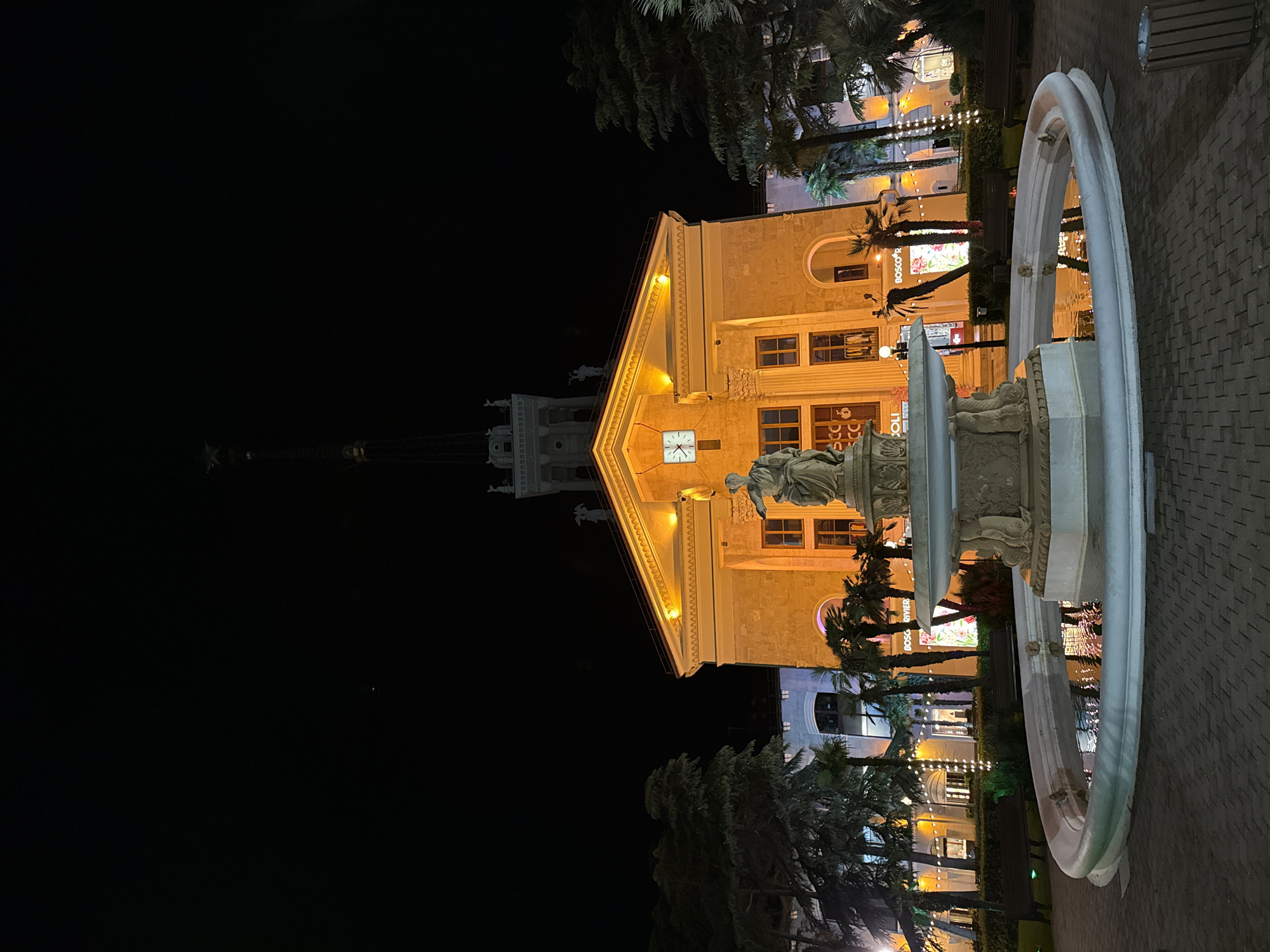 A nighttime exterior shot captures a grand building illuminated with warm orange light, featuring a prominent clock face and architectural details like columns and statues. In the foreground, a circular fountain with a statue at its center reflects the surrounding lights. Palm trees and other greenery are visible, along with benches and paving stones. The scene appears to be a public square or plaza. This is the Sochi Seaport (Sochi Morskoi Vokzal), a landmark in Sochi, Russia, known for its distinctive Stalinist architecture.