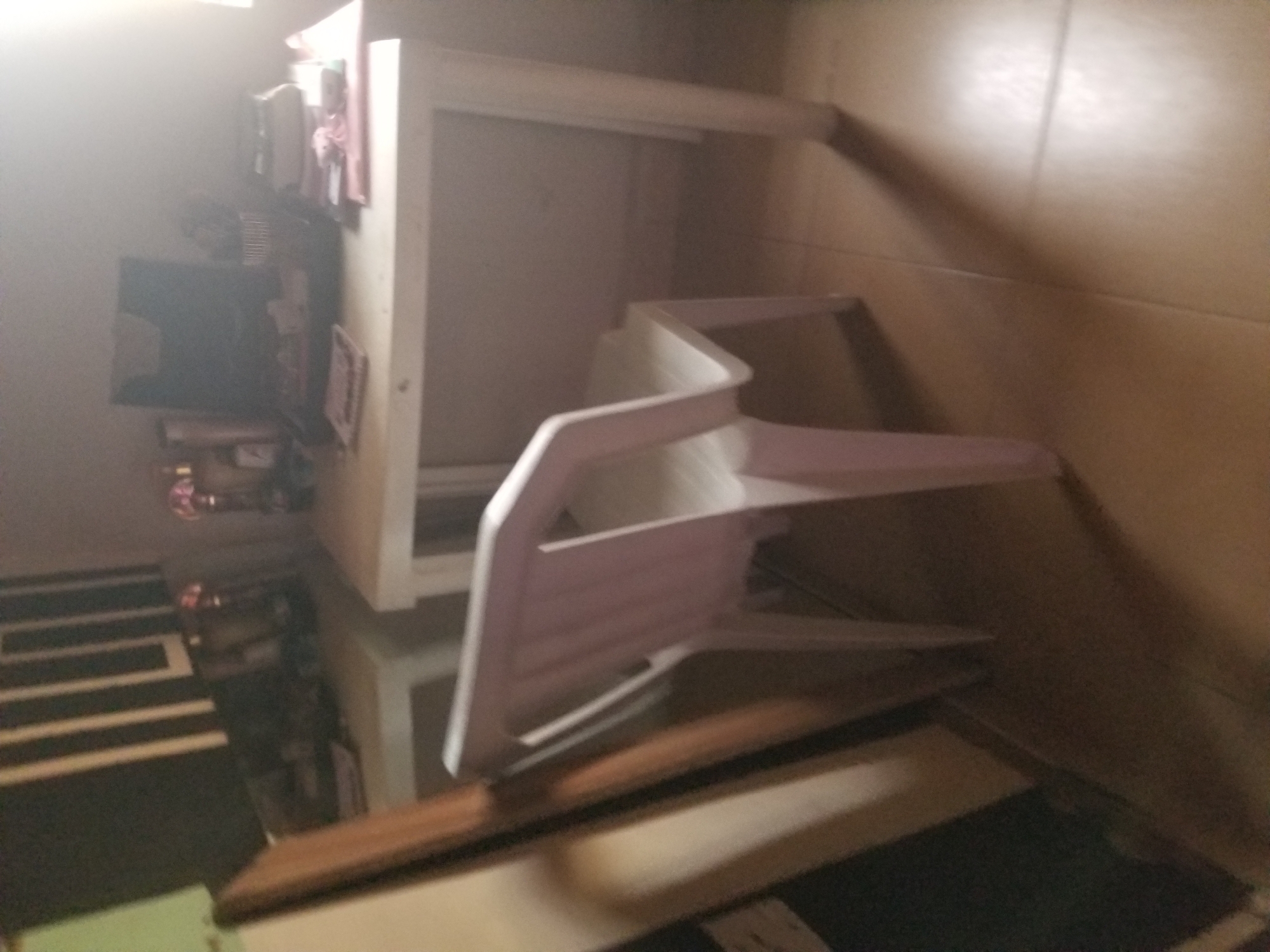 The image depicts an indoor scene, likely a room in Jauro Yinu, Nigeria, that appears to be a home office or study area. A white plastic chair is positioned in front of a white table. On the table, various items are present, including what seems to be a laptop or monitor, papers, and a pen holder. The background features a dark cabinet with vertical white stripes. The floor is tiled in a light brown color. The lighting suggests it is daytime, with a bright light source coming from the right side of the image. There are no people visible in the scene. The overall impression is one of a functional but somewhat cluttered workspace.